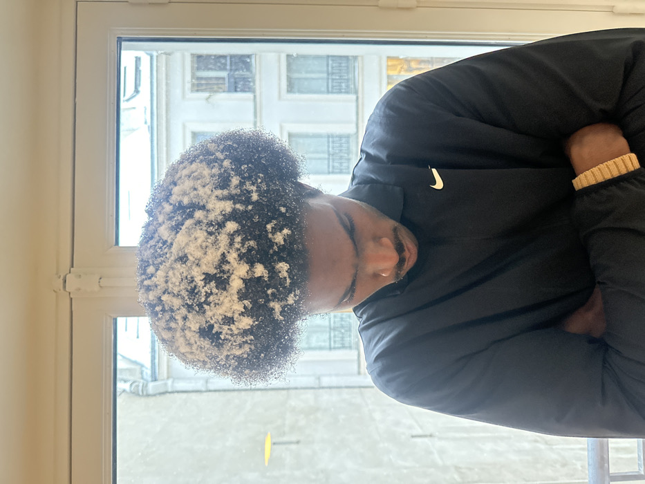 An individual, identified as a young Black male, is depicted from the mid-torso up, standing indoors in front of a window. His head is bowed, and his gaze is directed downwards. He possesses a dark, tightly coiled afro hairstyle, which is heavily covered with a white, powdery substance resembling snow or a similar particulate. He wears a black long-sleeved collared garment, possibly a jacket or shirt, featuring a white Nike swoosh logo on the left chest area. His arms are crossed over his chest, and a light brown, possibly woven or textured, bracelet is visible on his right wrist.
The background, visible through a white-framed window, shows the exterior of multi-story buildings characteristic of Parisian architecture, specifically Haussmannian style. These buildings are light-colored (off-white or pale grey) with symmetrically arranged windows and subtle decorative elements, indicative of a dense urban environment in Paris, France. The natural light suggests an overcast or bright day, with no direct sunlight. A faint yellow light source is visible in the distant left background through the window.