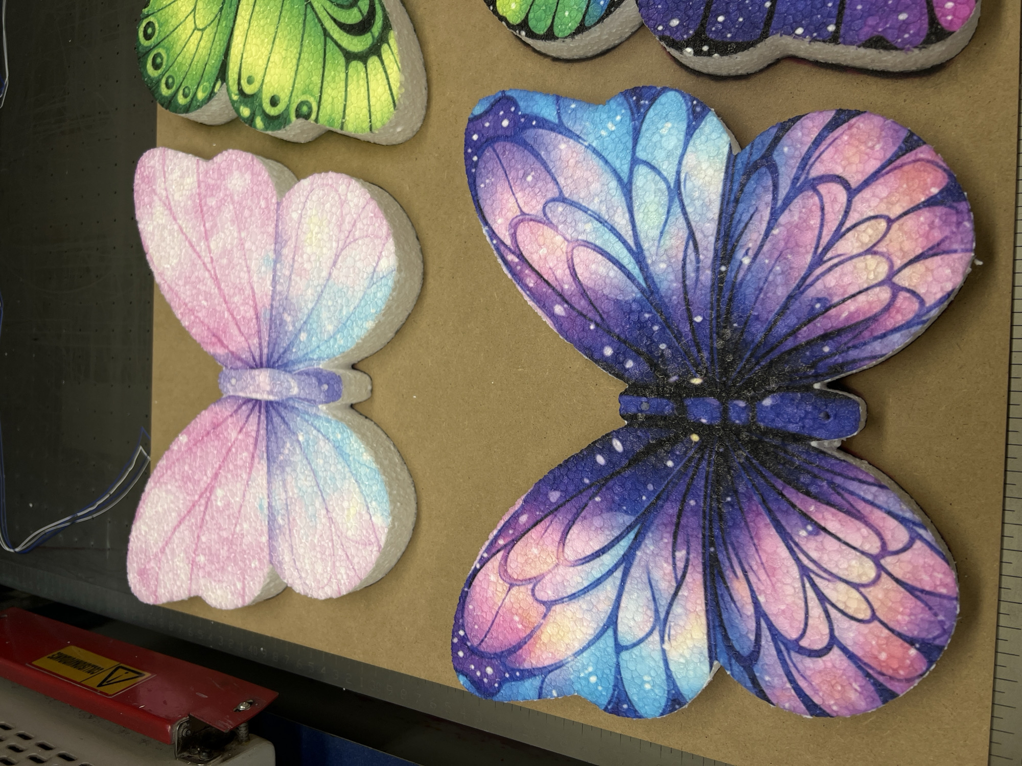 The image captures several colorful, butterfly-shaped cutouts displayed on a flat, light brown surface, likely wood or cardboard. These cutouts appear to be made from a thick, porous, foam-like material, possibly polystyrene, with detailed designs printed on their surfaces.
The most prominent cutout, located on the right, is a large butterfly featuring a vibrant "galaxy" or "starry night" design. Its wings transition through shades of dark purple, blue, and pink/orange, speckled with numerous small white dots resembling stars. Distinct black vein lines outline its wing patterns. To its left, a smaller butterfly exhibits a softer color gradient of pink, purple, and light blue, with less pronounced wing veins. Partial views of other cutouts are visible: a bright green butterfly at the top left with black veins, and a portion of another dark-colored butterfly at the top right.
The setting suggests an industrial or workshop environment. In the lower left quadrant, part of a machine or equipment is visible, including a red metal component. This component features a yellow warning triangle with a black exclamation mark inside, indicating caution. Faint, unreadable text is present beneath this symbol. White and blue electrical wires are also seen extending upwards from this area. Along the right edge of the brown surface, a ruler or measuring tape with visible numerical markings suggests precision or measurement is involved in the process. The overall scene implies these decorative items are either in production, being prepared, or arranged for display within a manufacturing or crafting context.