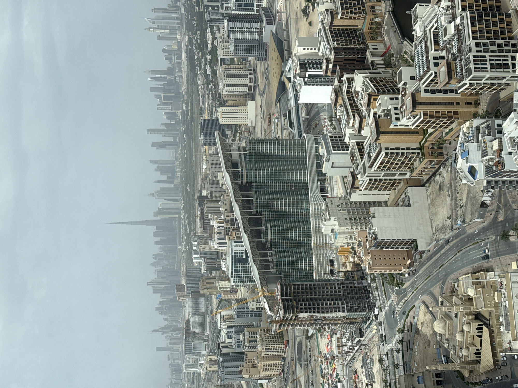 An aerial view of a densely populated urban landscape, dominated by numerous high-rise buildings, under a hazy sky. The architecture is modern, with a variety of shapes and styles evident in the construction of the buildings. Some structures appear to be under construction, with cranes visible amidst the building frameworks. Roads and traffic are visible amidst the buildings. The cityscape stretches into the distance, where the iconic Burj Khalifa is visible.