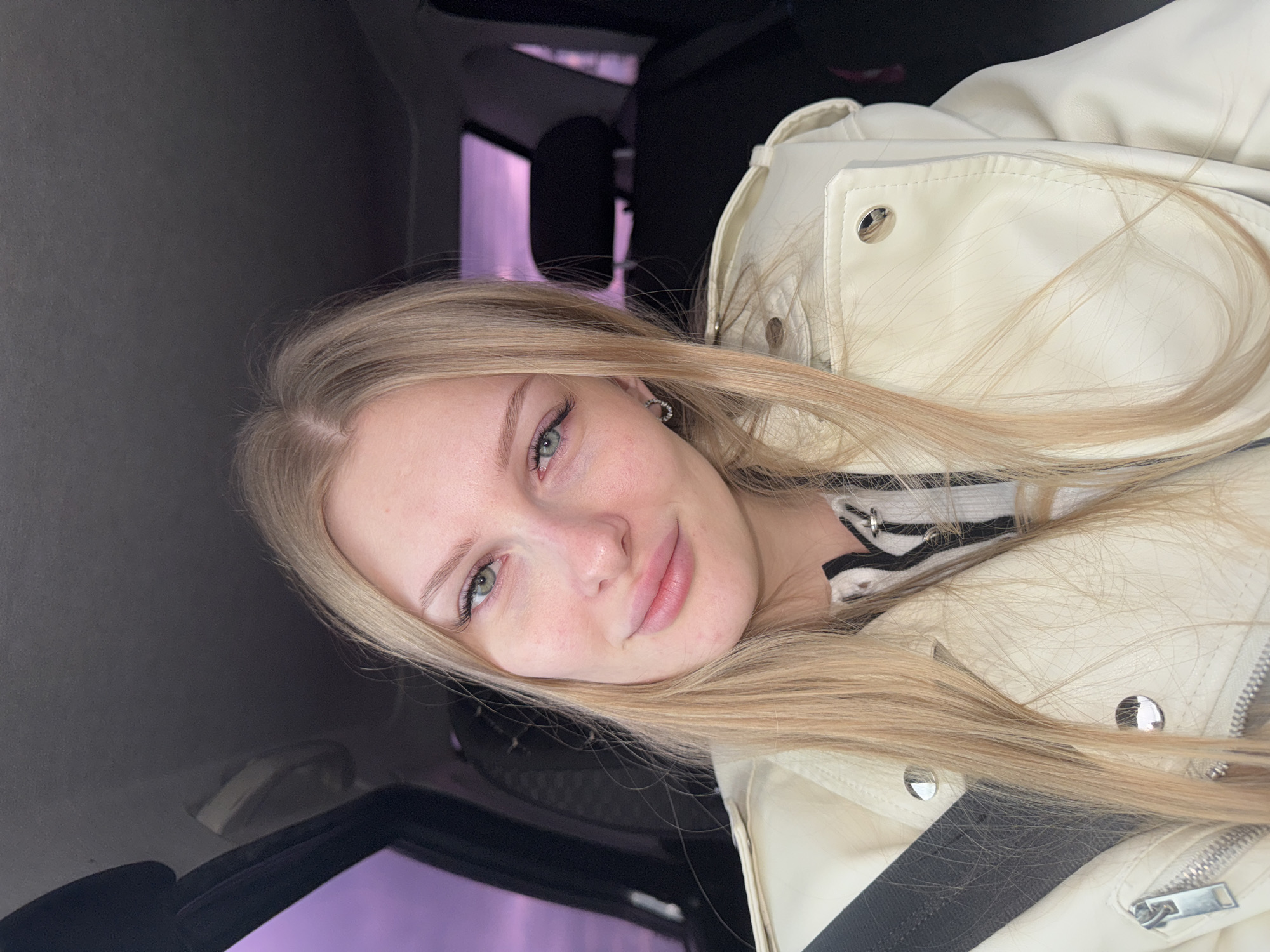 A fair-skinned young woman with long, blonde hair is inside a vehicle, possibly a car. She is wearing a light-colored jacket. The background shows the interior of the car and a glimpse of what may be outside the window. The location is assumed to be Perm, Russia.