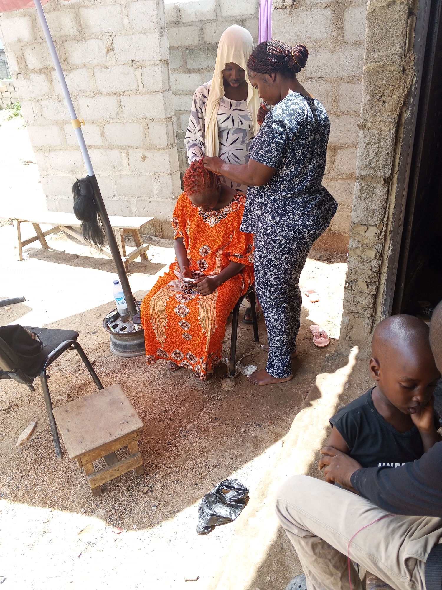 The image shows three women and a young boy in an outdoor setting in an unknown city and country. A woman in an orange floral dress is seated and looking at a phone. A woman with her hair in a bun, wearing a navy blue floral patterned jumpsuit, stands behind her and appears to be styling the seated woman's hair. A third woman in a white head covering and patterned top stands in the background, observing. To the right of the frame, a young boy in a black t-shirt is partially visible, seated next to a person in khaki pants. The surroundings consist of sandy ground, a partially constructed brick wall, a wooden stool, and miscellaneous items like a tire, bottle of water, and a black plastic bag. An umbrella pole is also visible, with a black hairpiece attached.