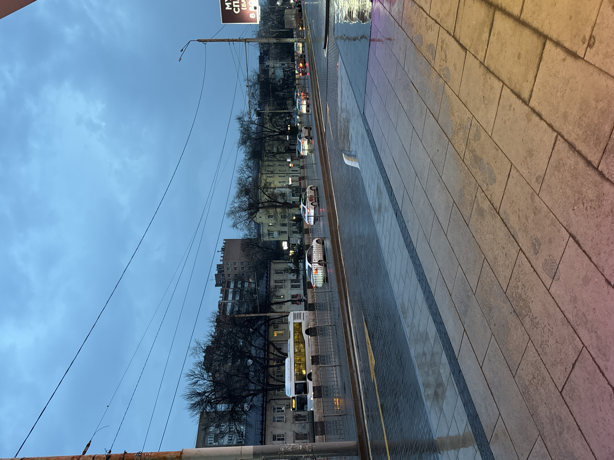 The image depicts a street scene in Ol'ginskaya, Russia during twilight or dusk. The perspective is from an elevated position looking down at an angle. The street is wet, likely from recent rain, with reflections visible on the paved surface. A sidewalk runs along the right side of the frame, made of large rectangular blocks. A bus and several cars are visible in the street. Buildings line the left side of the street, interspersed with bare trees. Overhead wires stretch across the sky, which is a muted blue-gray. A sign is visible in the upper left of the frame, with illuminated text in a Cyrillic script.