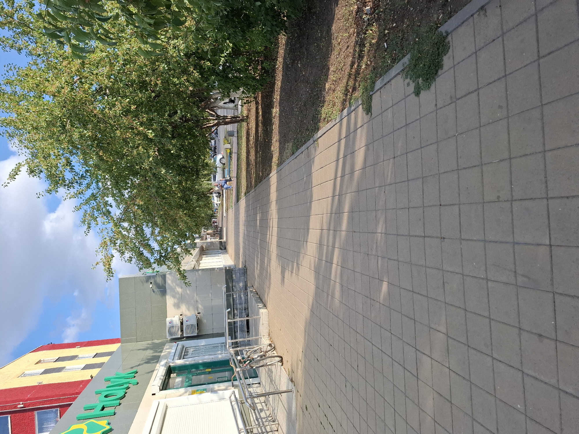 The image shows a sidewalk paved with square grey tiles in front of a building with a red and yellow facade on the left, and a row of trees on the right. A bicycle is parked on the sidewalk near a ramp. In the background, people are visible sitting on benches and cars are parked along a street. The sky is blue with scattered white clouds. The scene appears to be in an urban environment. The building on the left has a sign with green lettering that appears to say "Halyk". This suggests the location is likely a bank or financial institution. Given the information provided, the location is identified as the Halyk Bank branch in Lisakovsk, Kazakhstan.