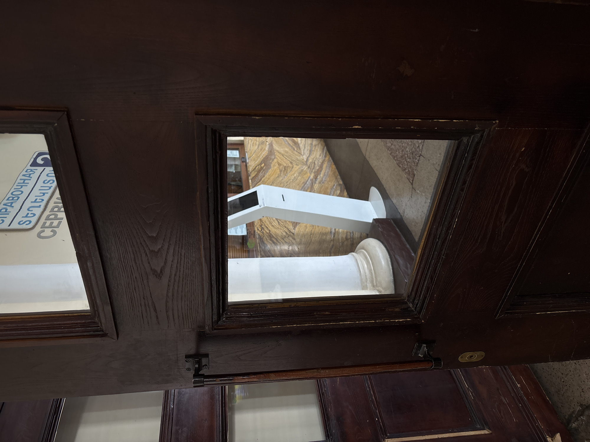 The image shows a dark brown wooden door with two glass panes. The upper pane reveals a sign with blue lettering that appears to be in Cyrillic script and Armenian script, with the word "СПРАВОЧНАЯ" (information) visible in Russian and "Տեղեկատու" (information) visible in Armenian. Below this, the word "СЕРВИС" is partially visible. The lower pane shows the reflection of a white kiosk or information terminal situated in a hallway with marble flooring and a patterned wall. A large white pillar is also visible. Based on the Armenian script on the sign, the scene is likely located in Yerevan, Armenia, possibly at a train station or a public information center. The door is open, with a black handle and latch visible on the left side. There are no people visible in the image.