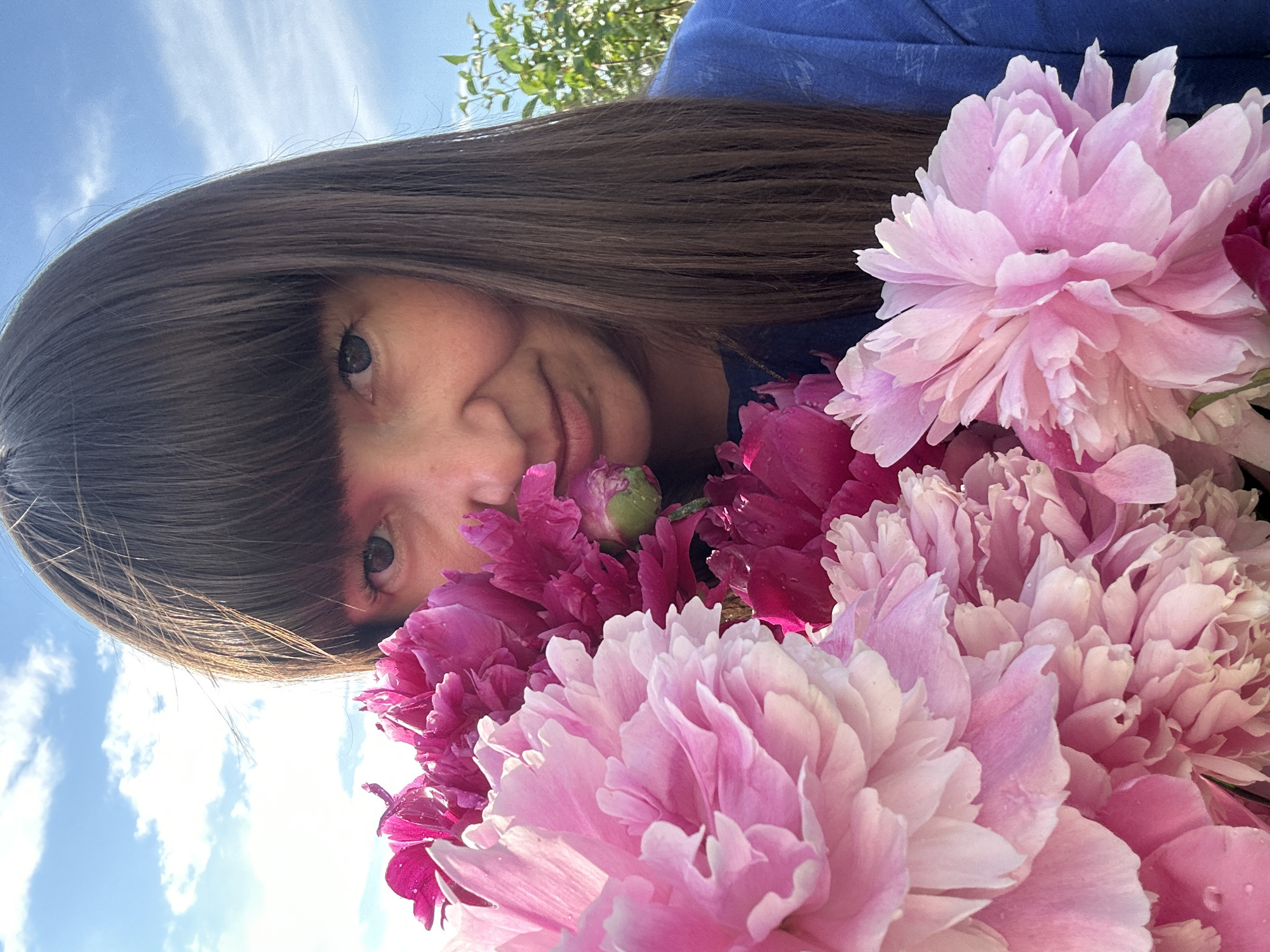 A woman with dark hair and bangs is holding a bouquet of pink and deep magenta peonies in front of her face. She is wearing a blue shirt and is outdoors with a blue sky and white clouds visible behind her. The peonies are large and have many petals. Some of the petals appear to have water droplets on them. The scene appears to be in a garden or outdoor setting. The location is described as "Среднее Кечево, Russia".