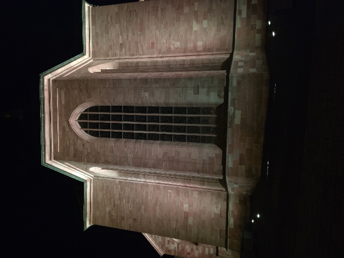 This nighttime photograph captures an exterior view of the Alexanderskirche in Zweibrücken, Germany. The building is constructed from reddish-brown sandstone blocks and is illuminated from below by multiple ground-level spotlights that cast an upward, warm glow onto the facade. The central feature is a tall, narrow, pointed-arch window, divided into numerous dark panes by a grid of mullions and transoms, indicating unlit interior spaces. Architectural elements resembling buttresses or recessed arched panels flank the main window. A distinct green-patinated copper trim or roofline outlines the upper edges of the structure, which presents a multi-faceted or polygonal form consistent with a church apse or chancel. The immediate foreground shows a dark, paved surface, with small, bright ground lights visible at the base of the building. The sky is completely dark, indicating a late-night or very early morning setting. No individuals, vehicles, or discernible actions are present within the frame.