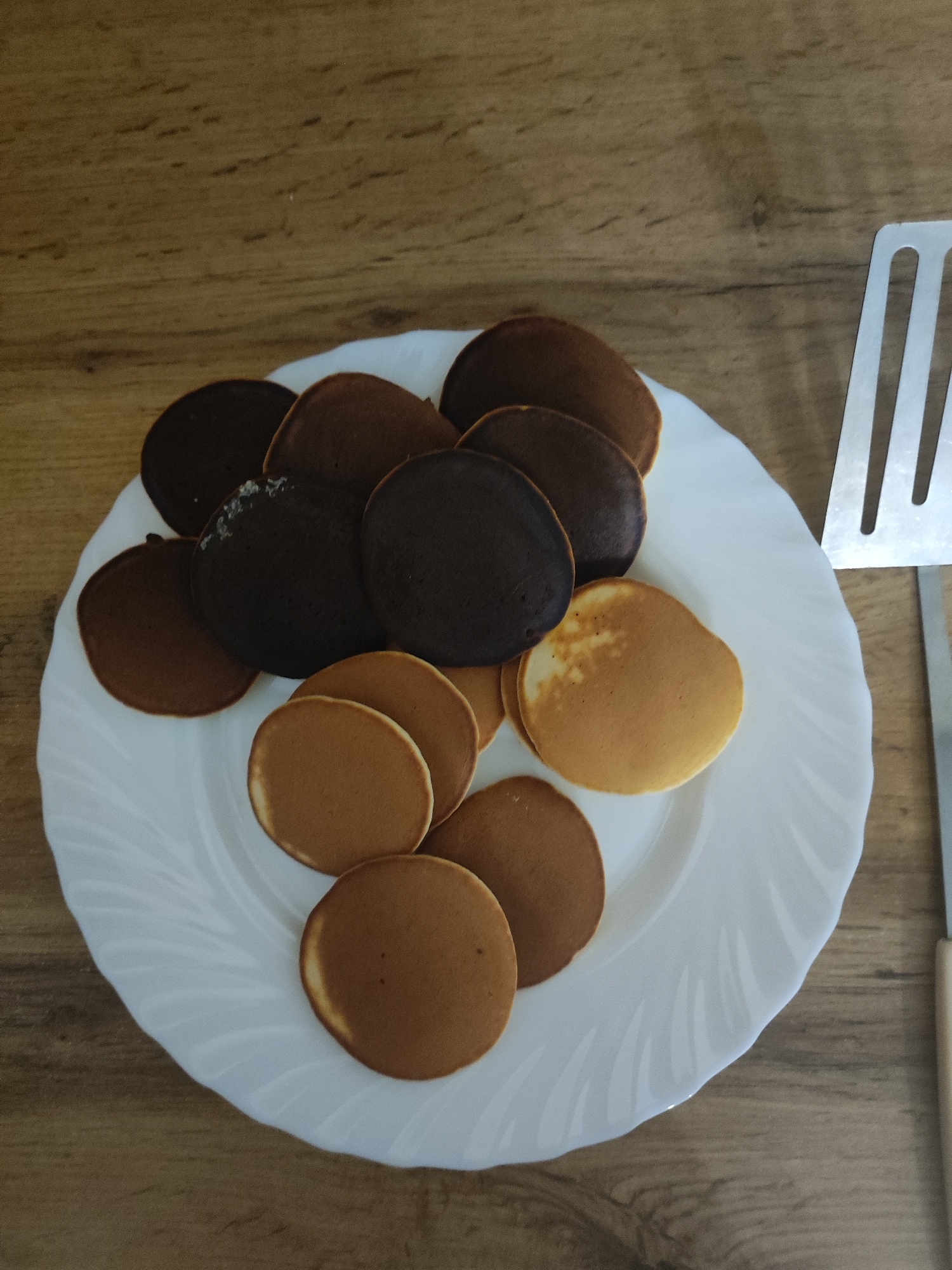 The image shows a white plate piled with small, round pancakes of varying shades, ranging from light golden brown to dark brown and nearly black. A metal spatula with a light-colored handle rests on the right side of the plate and table. The plate sits on a wooden surface. The location is not identifiable as Votkinsk, Russia based solely on the visual information provided.