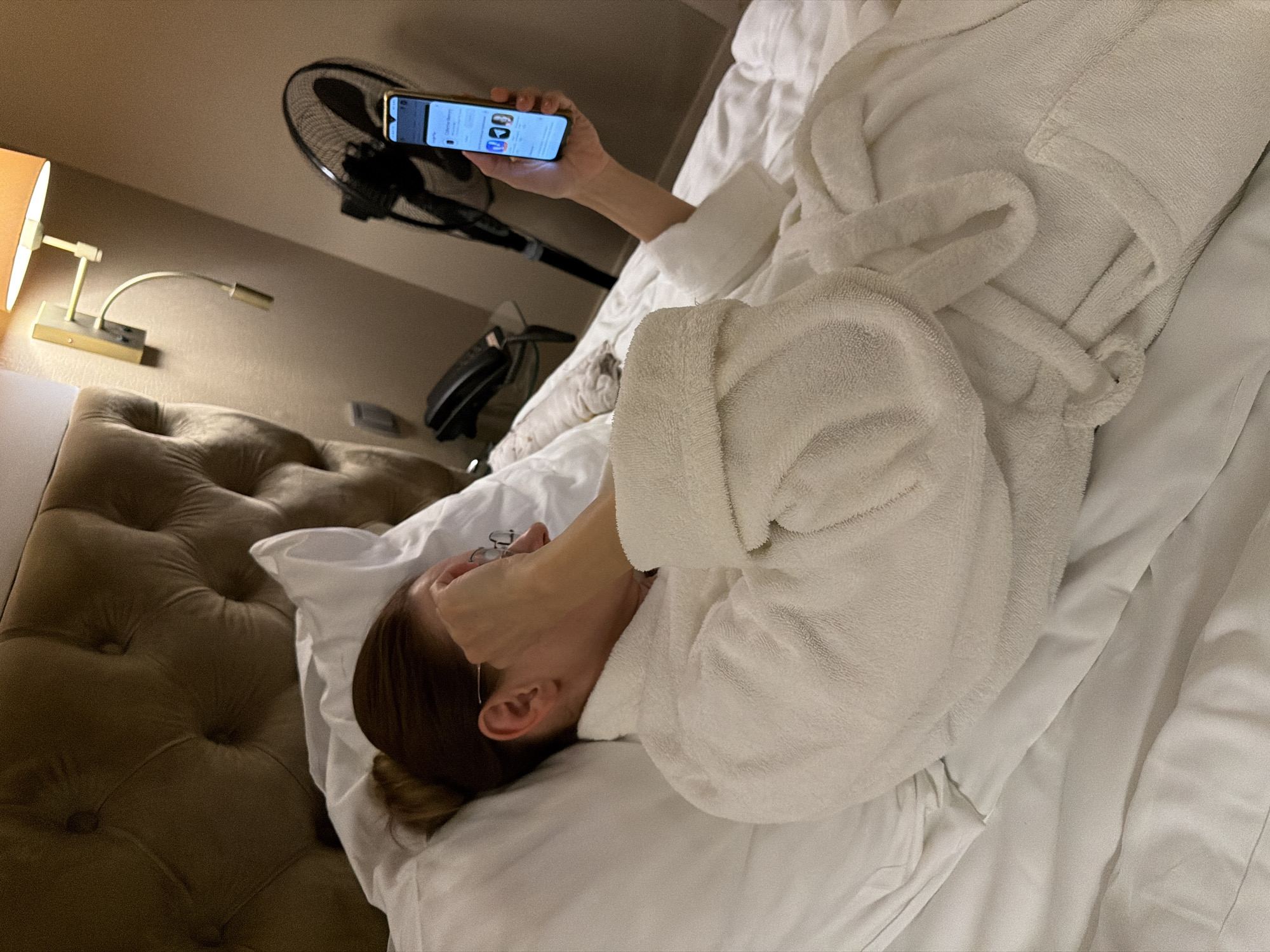 The image captures an adult female lying on a white bed in a hotel room in Svetlogorsk, Russia. She is wearing a white terrycloth bathrobe and has her hair pulled back into a bun. Her left hand is raised, partially covering her eyeglasses and the left side of her face. With her right hand, she holds a gold-colored smartphone vertically, its screen illuminated and displaying what appears to be a list of applications or products, indicated by three visible icons (two white, one black). The bed features a brown, tufted upholstered headboard. To the right of the subject, a wall-mounted lamp with an orange fabric shade provides illumination, accompanied by a smaller, flexible reading light arm. In the background, a black standing electric fan is visible, along with a black landline telephone resting on a surface. The walls of the room are a neutral tan color.