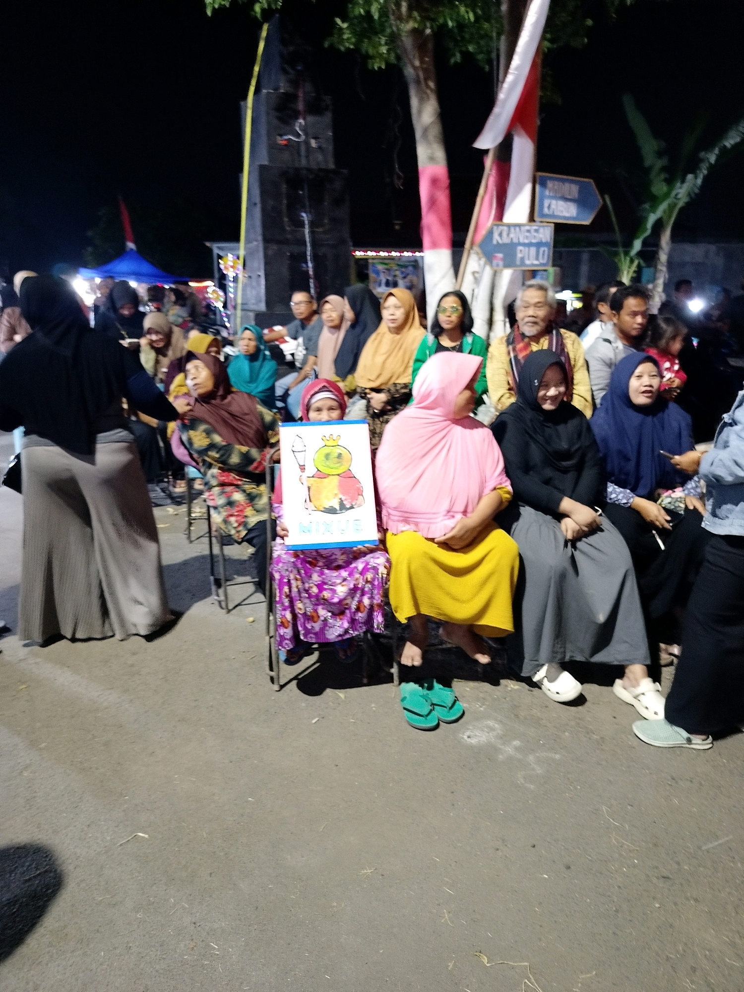 The image depicts an outdoor gathering at night, likely a local event or market in Putat, Indonesia. In the foreground, several people are seated on chairs. One woman in the center is holding up a drawing of a cartoon character with a crown, possibly a representation of a queen or a royal figure, with an ice cream cone next to it. The drawing is on a white poster board. She is wearing a purple patterned skirt and a red headscarf. To her left, a woman in a pink hijab and yellow dress is also seated. To her right, a woman in a black hijab and grey skirt is seated. Several other individuals are visible in the background, seated and standing. Behind them, a large dark structure, possibly a speaker or monument, is visible, adorned with red and white flags. Signs indicating directions are also present. The environment appears to be a paved area, suggesting a street or plaza. The lighting is dim, typical of nighttime events, with some artificial illumination present.