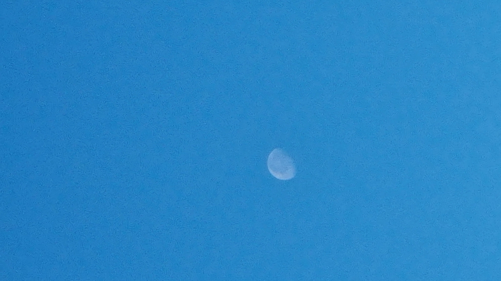 The image shows a crescent moon in a clear blue sky. The moon is positioned slightly to the left of the center of the frame and is illuminated from the right. The sky is a uniform, bright blue, suggesting it is daytime. There are no other celestial bodies or objects visible in the sky. The image is a vertical shot.