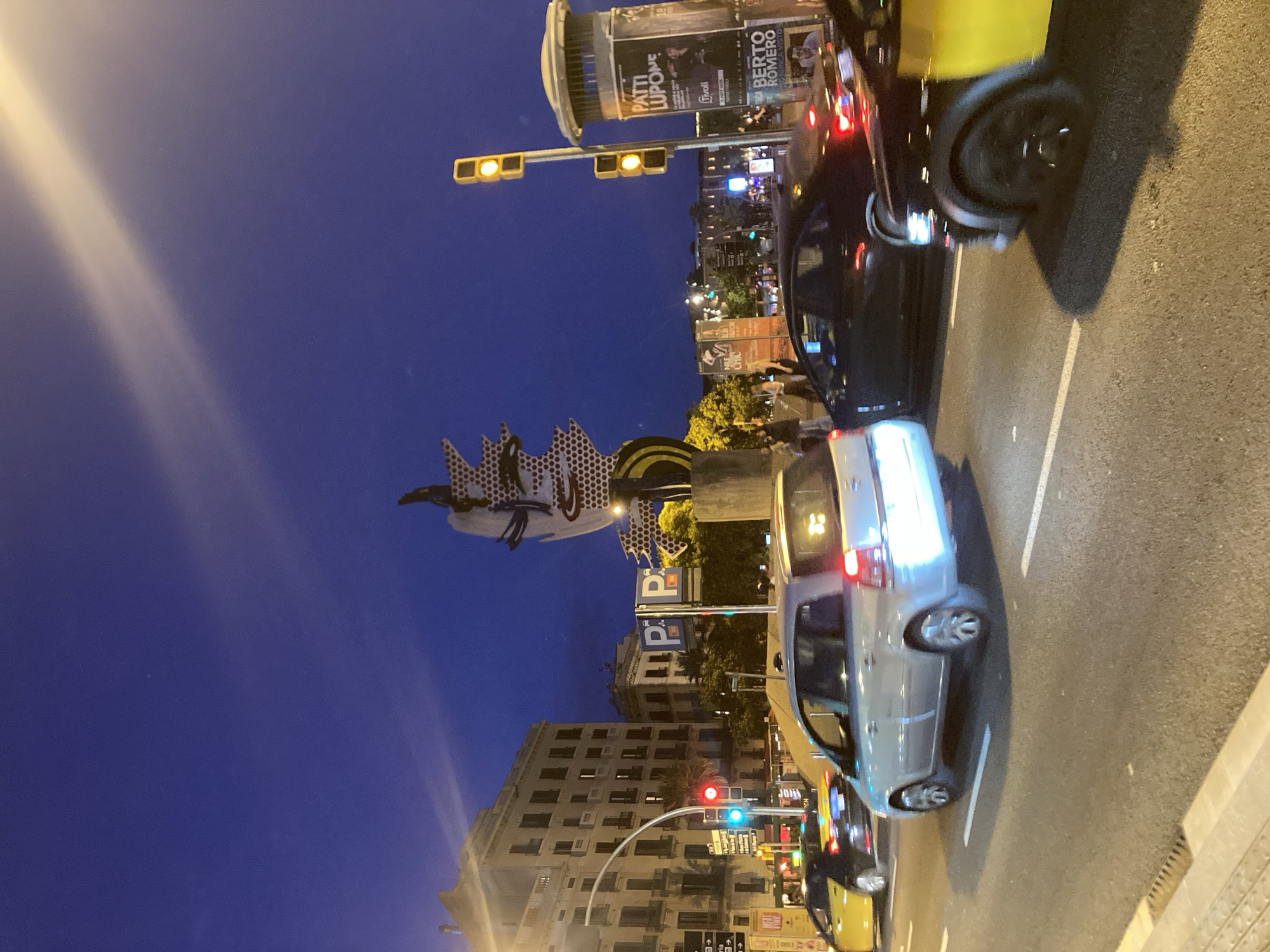 The image captures a street scene in Barcelona, Spain, at dusk or night. A prominent sculpture, resembling a stylized human face with pop art elements, stands in the background. The sculpture appears to be part of a public art installation or landmark. Several cars are on the road, indicating traffic. In the foreground, a silver minivan is in motion, its brake lights illuminated. A black sedan is behind it, also in motion. To the right, the side of a yellow taxi is visible. Traffic lights and signage are present, including what appear to be parking signs with the letter "P". Buildings line the street, featuring multiple stories and windows. The sky is a deep blue, transitioning from daylight. There are streetlights and illuminated signs adding to the ambient light of the urban environment. Several pedestrians are visible on a sidewalk in the mid-ground, near the sculpture. The overall scene depicts a busy urban setting at night with a blend of traffic, architecture, and public art.