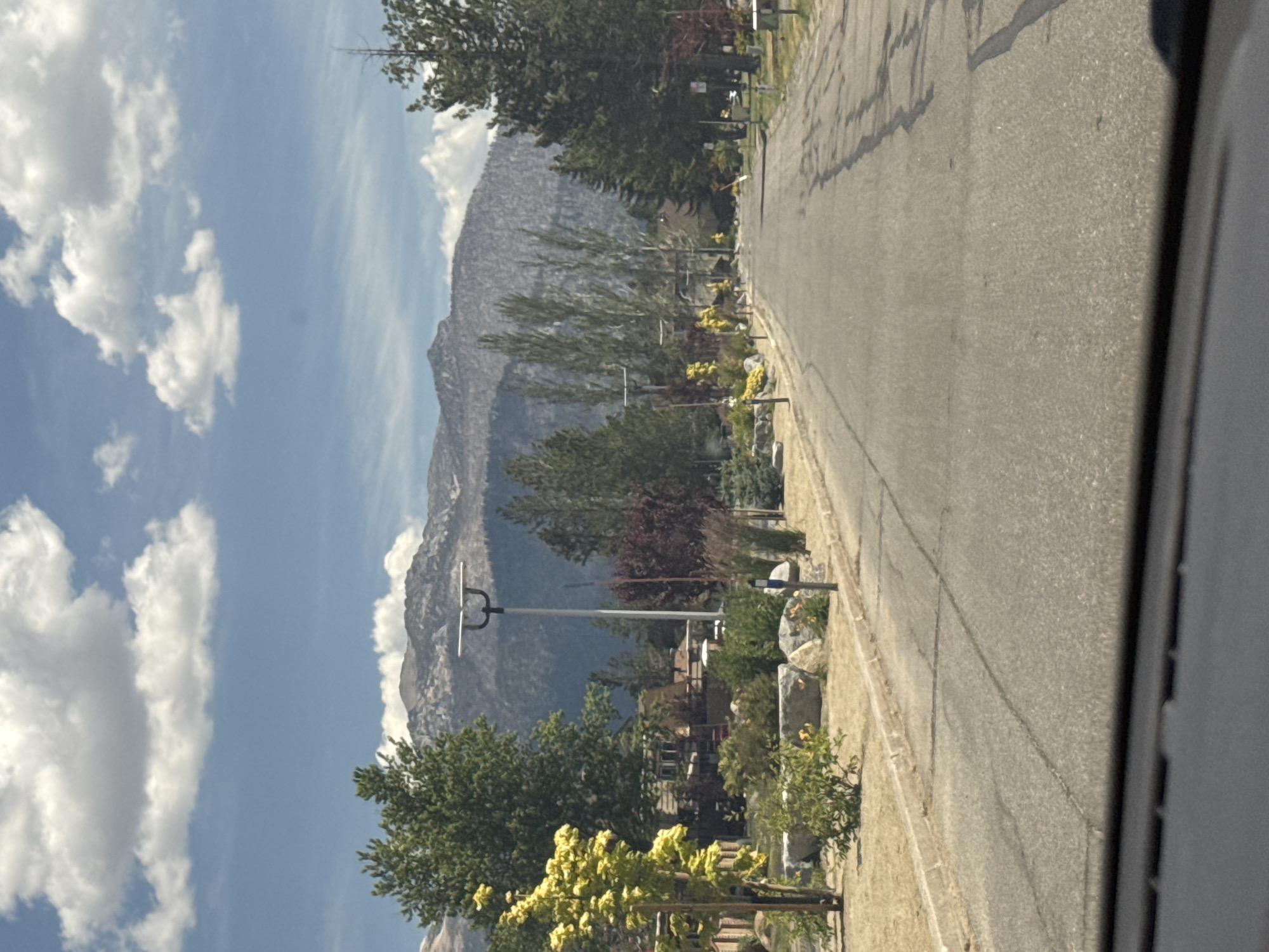 This is a medium-distance, eye-level, slightly downward-angled, outdoor, daytime photograph of a paved residential street in South Lake Tahoe, California, United States of America. The street is lined with manicured landscaping, including trees, shrubs, and rocks. In the distance, mountains are visible, covered with a light dusting of snow and trees. The sky is blue with scattered white clouds. The photograph appears to have been taken from inside a vehicle, as indicated by the dashboard in the foreground. The street is empty, with no people or vehicles visible. There are streetlights with solar panels along the road.