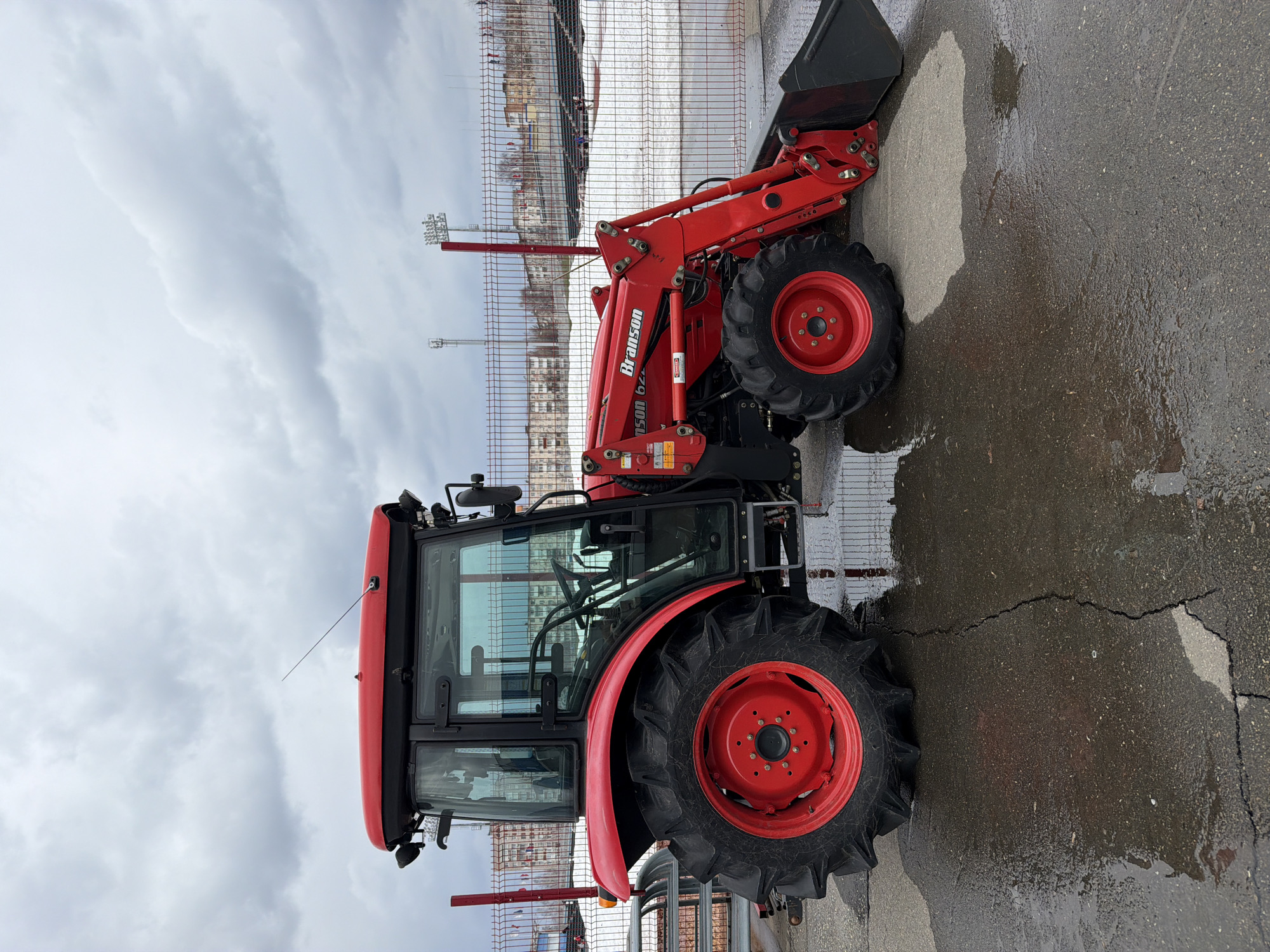The image shows a red Branson 6225C tractor with a front-end loader parked on a wet, cracked asphalt surface. Behind the tractor is a chain-link fence and what appears to be a stadium or sports complex with floodlights. Based on the details of the stadium, this photo may be in Votkinsk, Russia. The sky is overcast.