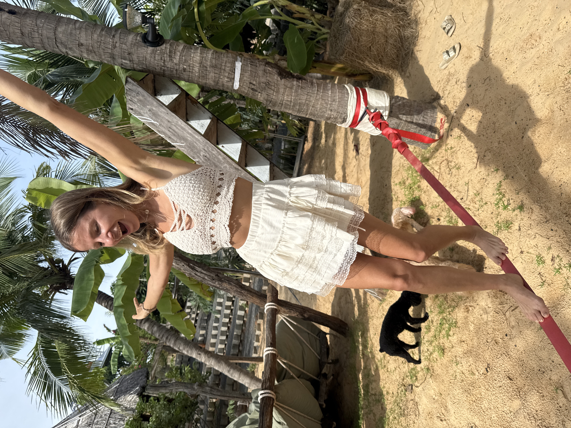 A blonde woman is walking on a red slackline in Mũi Né, Vietnam. She is wearing a white crochet crop top and a white ruffled skirt. Her arms are extended for balance. The ground is sandy and there are a few small green plants. In the background, there are palm trees, a wooden structure with stairs, and what appears to be a hut. Two dogs, one black and one light brown, are present on the sand. The woman is looking to her left with her mouth open, appearing excited or surprised. A straw bale and a pair of sandals are visible to the right.