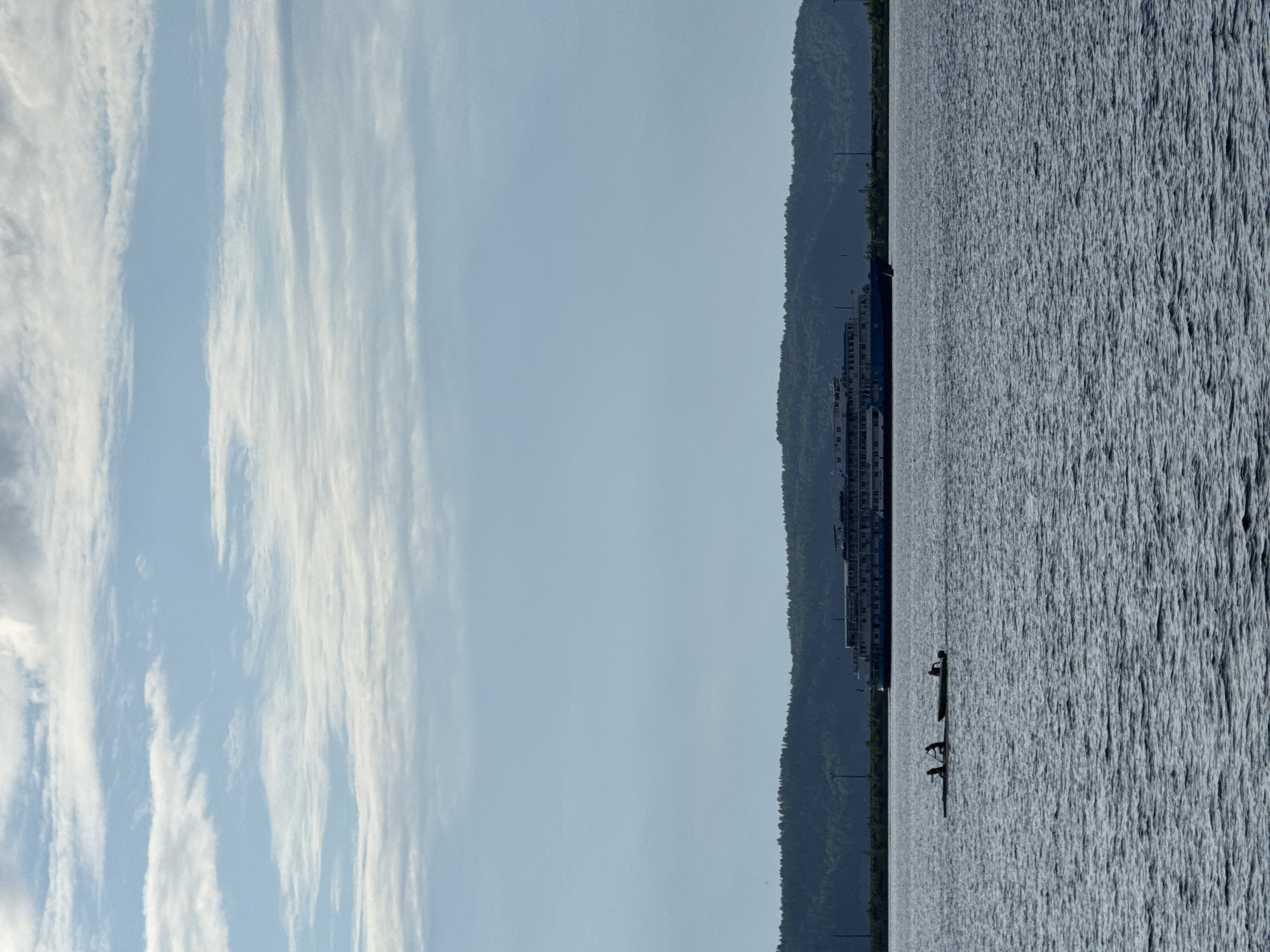The image captures a wide shot of the Kama River in Chaikovsky, Russia, under a sky filled with scattered clouds. In the foreground, the dark blue water is rippled by a gentle breeze. Two small boats with silhouetted figures are visible on the water; one appears to be a kayak with two occupants, and the other a small motorboat with one occupant. In the mid-ground, a large, multi-decked passenger ship is docked or cruising along the river. The ship has a white superstructure with blue accents and numerous windows. Behind the ship, a dense, dark green treeline forms the horizon, suggesting a forested hillside. A few utility poles are faintly visible in the distance along the shoreline. The overall scene depicts a tranquil day on the river, with human activity on a small scale contrasted against the vastness of the water, the imposing ship, and the natural landscape.