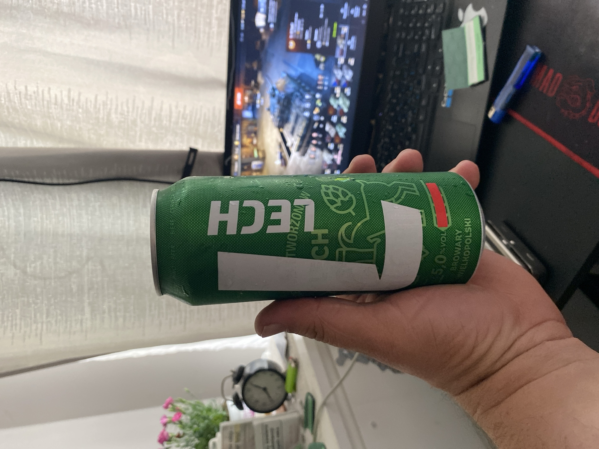 A hand holds a green can of Lech beer in front of a laptop displaying a video game with tanks. The background includes a sheer curtain and a windowsill with a desk clock and a potted plant. The scene is indoors, likely in a residential setting in Szczecin, Poland.