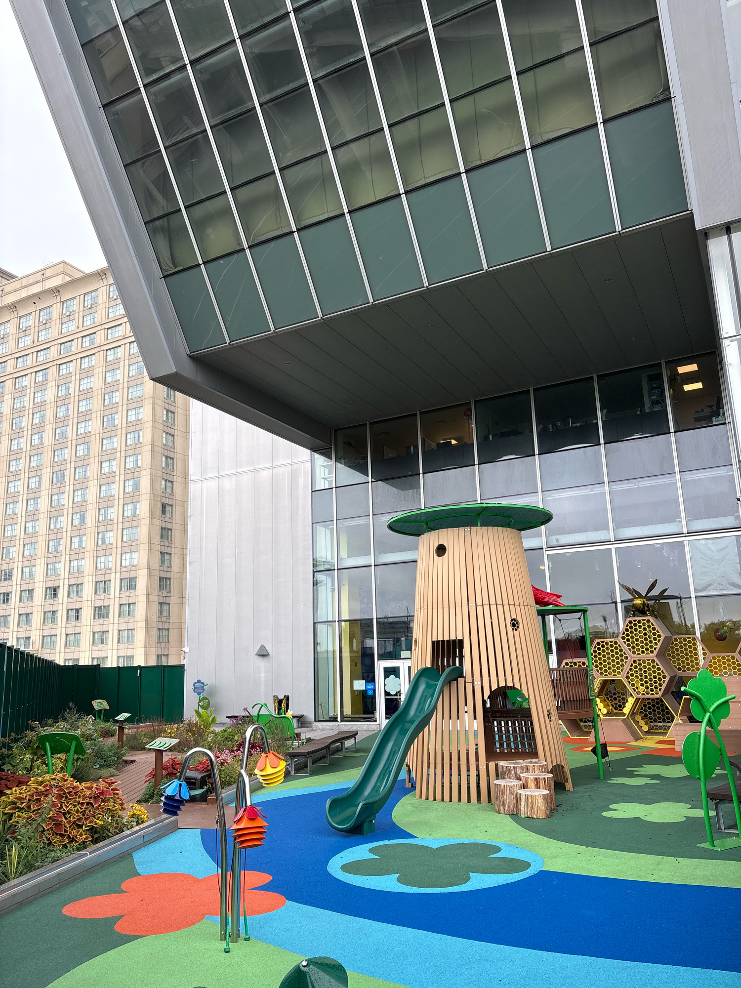 The image shows a modern playground located on a rooftop in Hartford, Connecticut, USA. In the background, a tall, beige building with many windows dominates the skyline. Above the playground, a sleek, modern building with a facade of glass windows and metal accents juts out over the play area. The playground itself features a colorful, impact-absorbing surface with blue, green, and orange patterns.
Central to the playground is a whimsical, treehouse-like structure made of light-colored wood with a green circular roof. A green slide emerges from this structure. Adjacent to the treehouse is a climbing structure resembling a honeycomb, with yellow hexagonal openings. There are also swings and other play equipment.
The foreground of the playground shows a section with bright green, blue, and orange surfaces, including flower-shaped cutouts. Several metal structures with colorful, tiered elements that resemble flowers are also visible. To the left, a neatly landscaped area with various plants and low green fencing adds a touch of nature to the urban setting. A green metal fence runs along the edge of the rooftop garden. The sky is overcast. No people are present in the image.