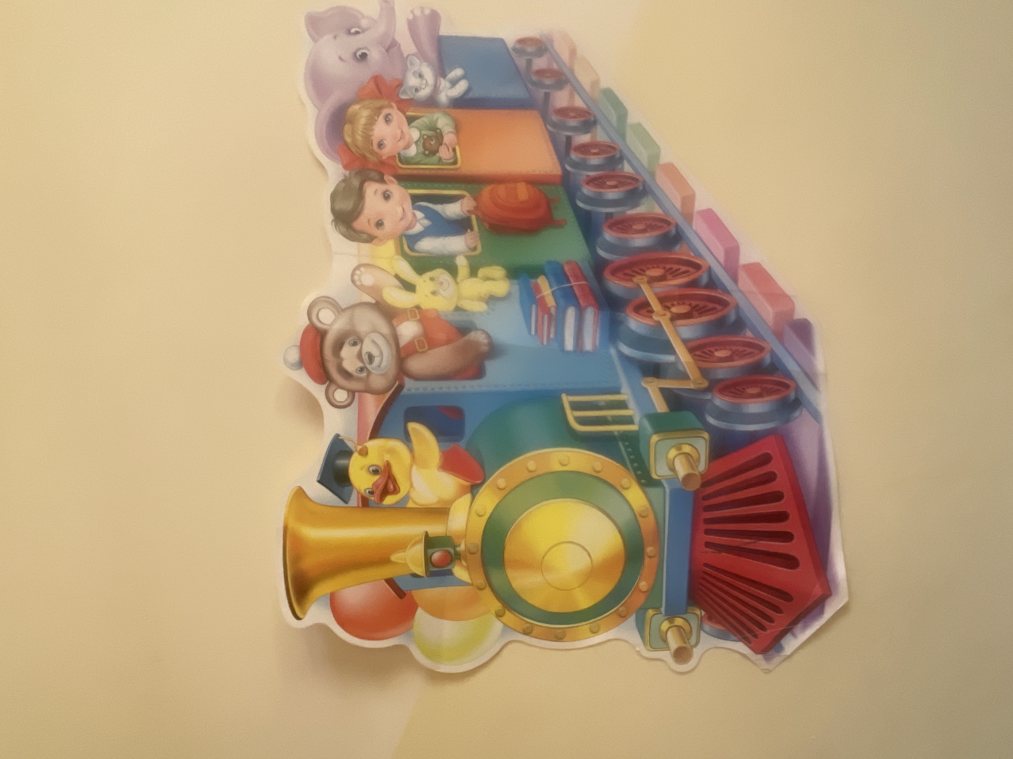 The image shows a colorful, cartoon-style depiction of a train filled with various characters. A yellow duck wearing a graduation cap and holding a red balloon is at the front of the train. Next to the duck is a brown teddy bear with a red hat, and a yellow bunny. Two children, a boy and a girl, are riding in the middle section of the train. The boy is wearing a blue vest, and the girl is holding a teddy bear. Behind them, a light purple elephant peeks out. To the right of the girl, a white cat is also visible. The train cars are stacked with colorful books, and the wheels are red. The train is depicted as if it is moving along train tracks made of colorful blocks. The overall scene is cheerful and whimsical. The background is a plain, light yellow wall. There is no visible indication of the city of Sochi, Russia, or any specific urban environment.
