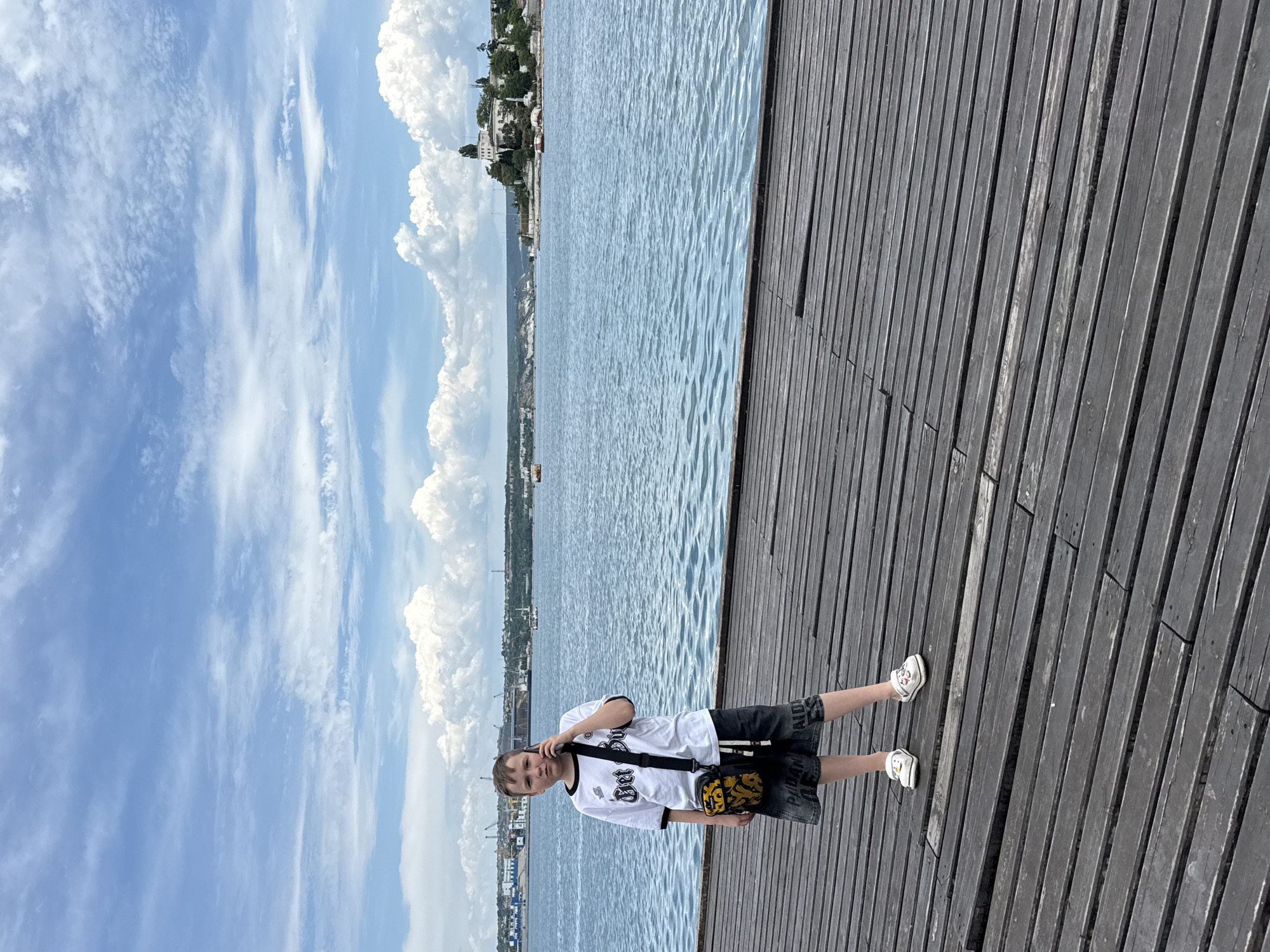 A young male, estimated to be between 10 and 14 years old, stands on a dark wooden pier. He is wearing a white short-sleeved t-shirt with black trim around the neck and sleeves, featuring a black graphic on the front. He has dark grey denim shorts with faint white lettering on the legs and white closed-toe sandals adorned with colorful cartoon faces. A black shoulder bag with a yellow and black patterned pouch is slung across his chest. He holds a dark-colored smartphone to his right ear with his right hand, suggesting he is on a call, and looks directly at the camera with a neutral expression.
Behind the boy is a wide expanse of blue-grey water with small ripples. In the distant background, the shoreline of a city or town is visible. To the left, there is an industrial area with various buildings, cranes, and structures along the water. To the right, a greener, hilly area features a prominent white building with a tower, surrounded by trees. The sky above is bright blue with numerous white cumulus clouds, and some wispy clouds are also present. The scene is illuminated by natural daylight.