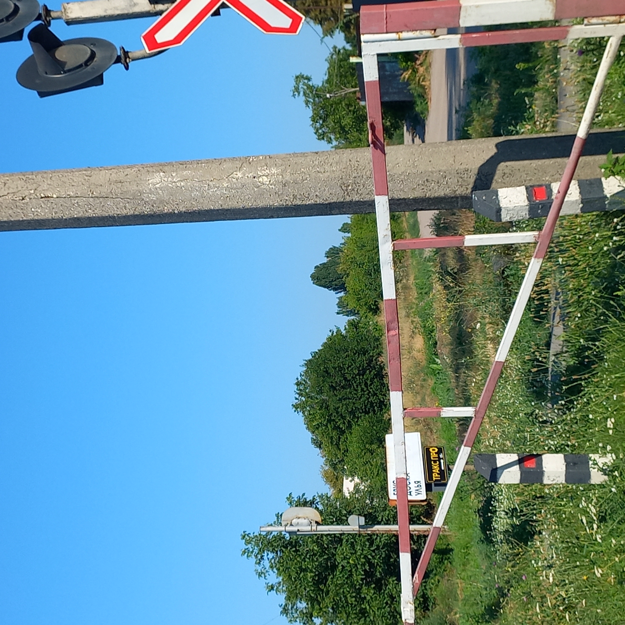 The image shows a railway crossing barrier in Kropyvnytskyi, Ukraine. The barrier is red and white striped, and it is in the lowered position, indicating that a train is likely approaching or passing. There is a traffic light on a pole to the right of the barrier, which appears to be dark. A signpost with two signs is visible behind the barrier. One sign is white with black lettering that reads "ДОСКА УЛЬЯ", and another smaller yellow sign below it reads "ТРАКС ПРО". The crossing is surrounded by overgrown grass and some trees in the background. The sky is clear and blue. The image quality is moderate, with some graininess.