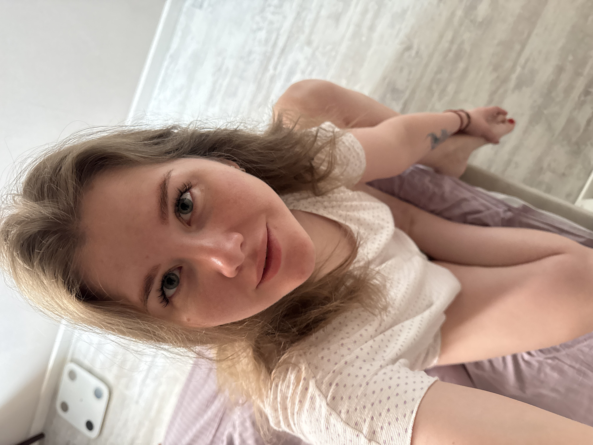 A young woman is captured in a close-up, slightly overhead selfie, appearing to recline or lie down in an indoor setting. She has long, wavy, light brown or blonde hair and light-colored eyes, which are directed at the camera with a gentle, relaxed smile. She wears a light-colored top with a subtle pattern of small, light purple dots.
Her body posture shows her legs bent, with her right foot resting comfortably, displaying red-painted toenails. Multiple thin, dark-colored bracelets adorn her right wrist, and a small, faded tattoo, possibly depicting a bird or abstract design, is visible on her right forearm.
The background includes a purple or lilac-colored sheet or duvet, suggesting a bed or sofa. On the light-colored floor in the lower-left, a white bathroom scale with four dark circular pads is visible. The lighting is bright and diffused, indicative of daytime. The overall atmosphere conveyed is relaxed and casual. There is no visible text in the image. The location context provided is Gornyi (Dvurechenskoe S/P), Russia.