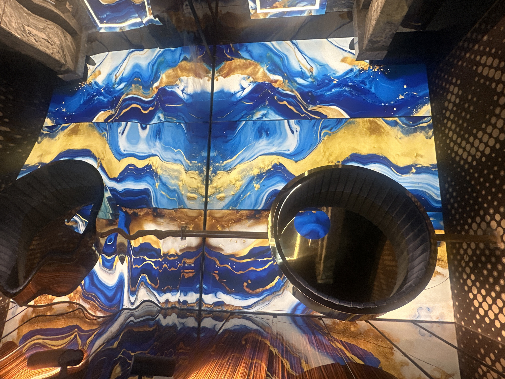 The image depicts an indoor scene with abstract, marbled artwork on the walls. The artwork features swirling patterns of blue, gold, and white. A spherical, dark-colored object with a metallic rim is suspended from the ceiling by a thin rod. A blue sphere is visible inside the larger sphere. The floor has a dotted pattern. The lighting in the scene is warm and reflective, casting shadows and highlights on the surfaces. The overall impression is one of modern, artistic interior design. There are no people or specific landmarks visible to indicate a city.