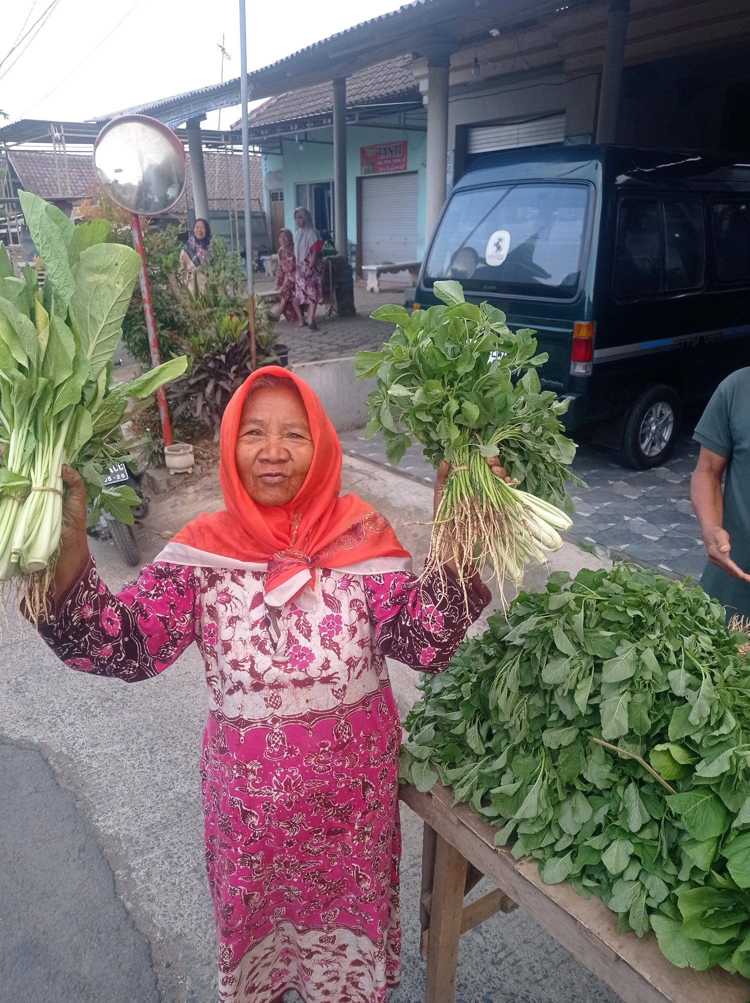 An elderly woman, likely a vendor, stands in front of a wooden stall laden with leafy green vegetables. She is wearing a vibrant pink and white patterned dress and an orange headscarf. In her left hand, she holds a bunch of green leafy vegetables with white roots, and in her right hand, another bunch of similar vegetables. To her right, a large pile of leafy greens rests on the wooden stall. Behind the woman, a dark green van is parked. In the background, a building with a corrugated roof and a blue facade is visible. The scene appears to be set outdoors on a street in Purwosekar, Indonesia, likely at a local market. The overall atmosphere suggests a typical day of commerce in a Indonesian village or town.