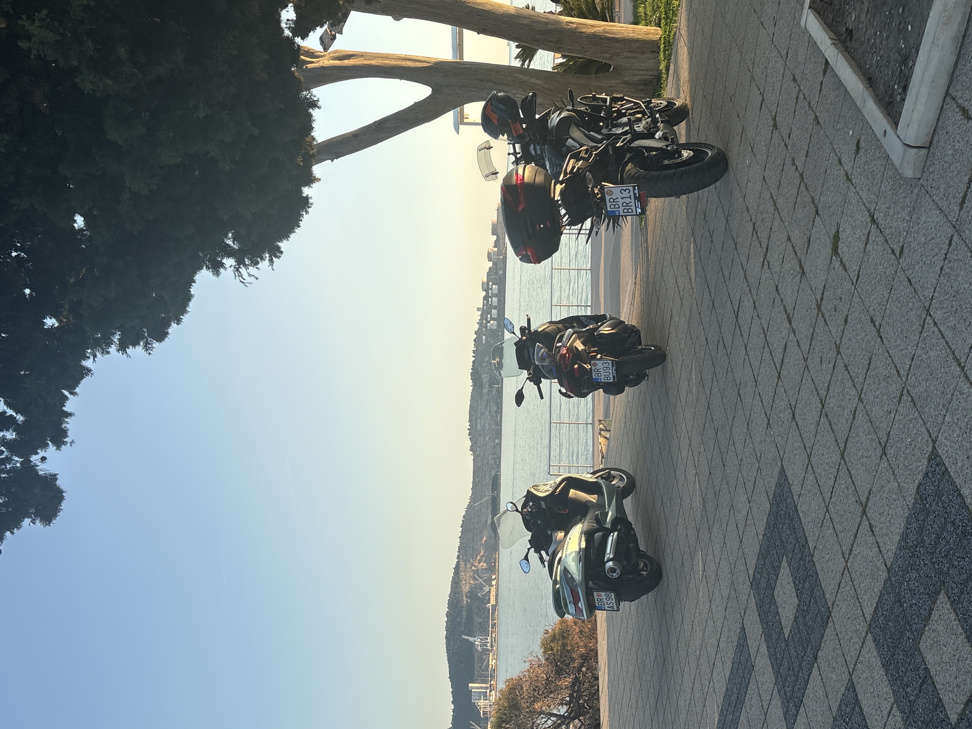 The image depicts a row of three motorcycles parked on a patterned sidewalk near the coast. The motorcycles, arranged in a line, have license plates with the prefix "BR", which identifies them as registered in Bar, Montenegro. Behind the motorcycles is a view of the Adriatic Sea, with a coastline visible in the distance. The foreground includes a tree and planter. The sky is clear with a hazy quality near the horizon. The image is taken during the day, likely during the early morning or late afternoon given the low angle of the light.