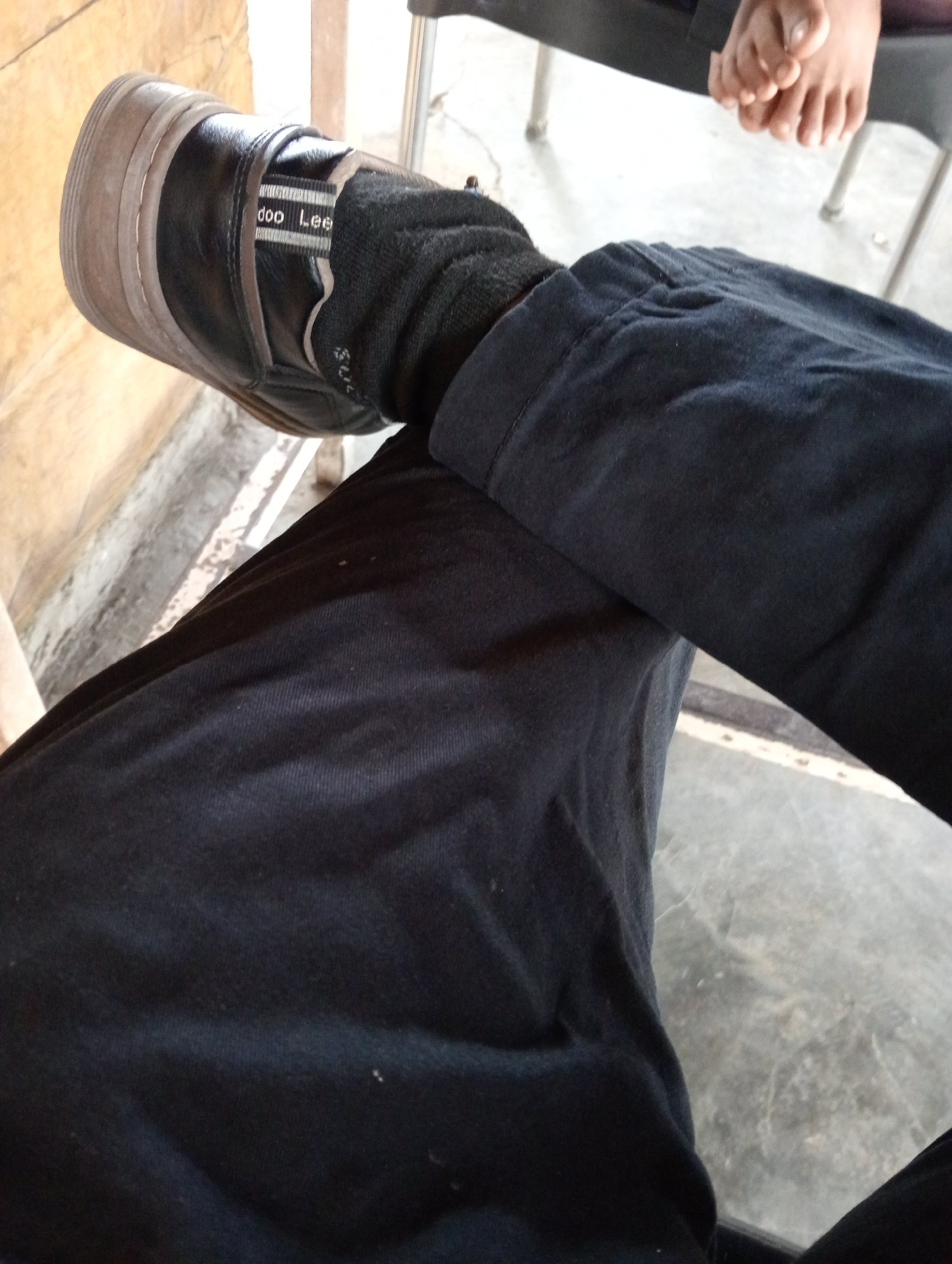 The image depicts a person sitting with legs crossed. They are wearing black pants, a black sock, and a black shoe with a thick sole. The shoe has a gray stripe with the name "Lee" on it. In the upper right corner of the image, bare feet are visible, belonging to another individual. The scene appears to be indoors, on a worn surface. The specific location in Lhokseumawe, Indonesia is unknown.