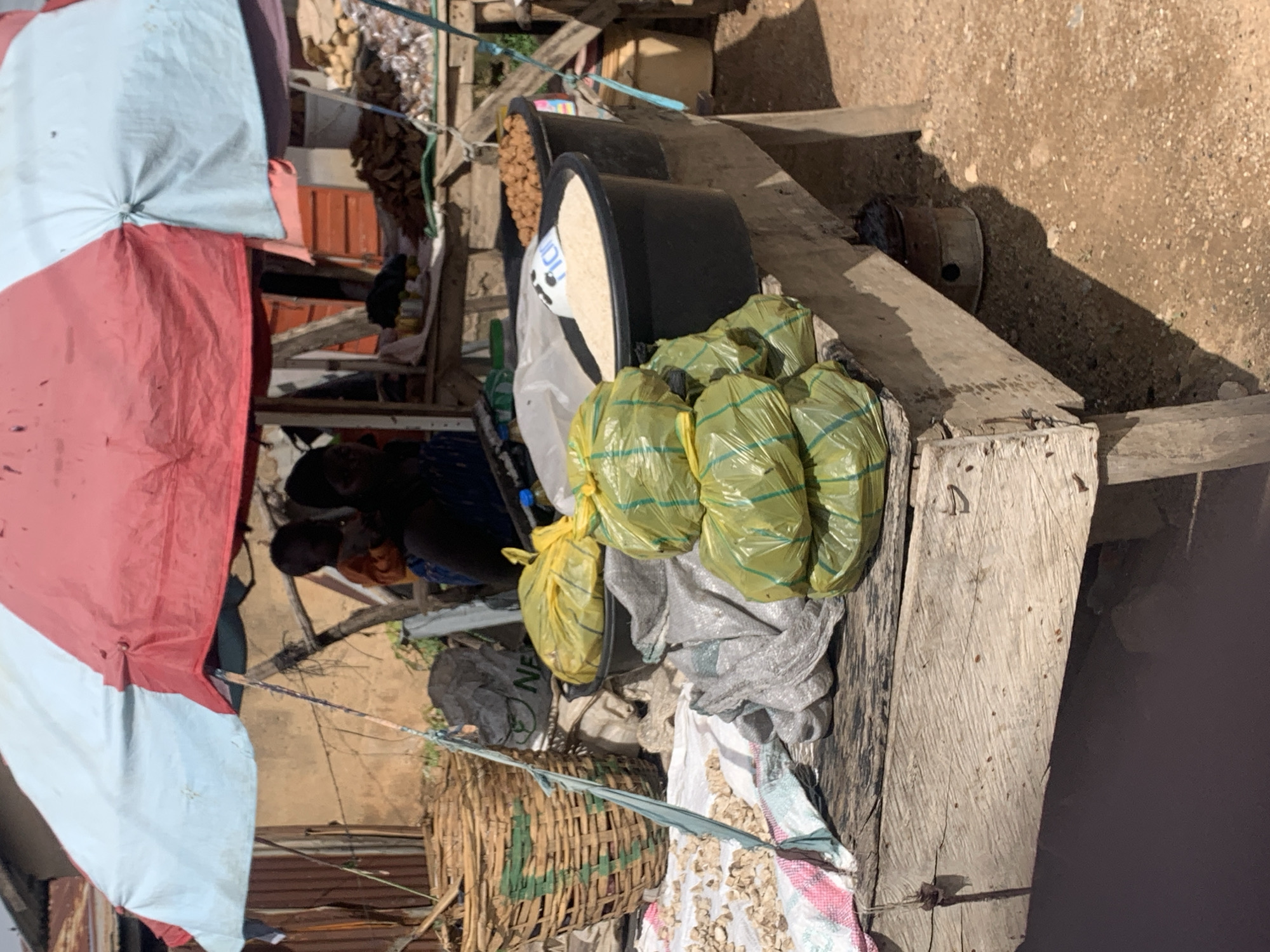 This is a medium shot of an outdoor market stall, likely in Jos, Nigeria, with a red and light blue striped umbrella providing shade. A weathered wooden table displays various goods. To the left, a large woven basket contains produce. In front of the basket, on a white and pink striped cloth, are scattered small, light-colored pieces of what appear to be dried roots or tubers.
Dominating the center of the image are stacks of several large, clear plastic bags filled with a yellow substance, possibly grains or dried goods, tied shut with yellow twine. Behind these bags are a black bucket containing white granular material, possibly rice, and another bucket filled with small, brown, spherical items, possibly nuts or beans. A partial white label with blue lettering that reads "ric" is visible on the edge of the rice bucket.
To the far left, partially obscured by the basket, is a crumpled gray plastic bag with green lettering. Behind the table, two individuals are visible. One, in the background, appears to be a child wearing an orange garment. Another person, in the mid-ground, is wearing a blue and white patterned top. The environment is dusty and the ground is covered in sand and small stones. A small metal object, possibly a stove, is visible beneath the table on the right.