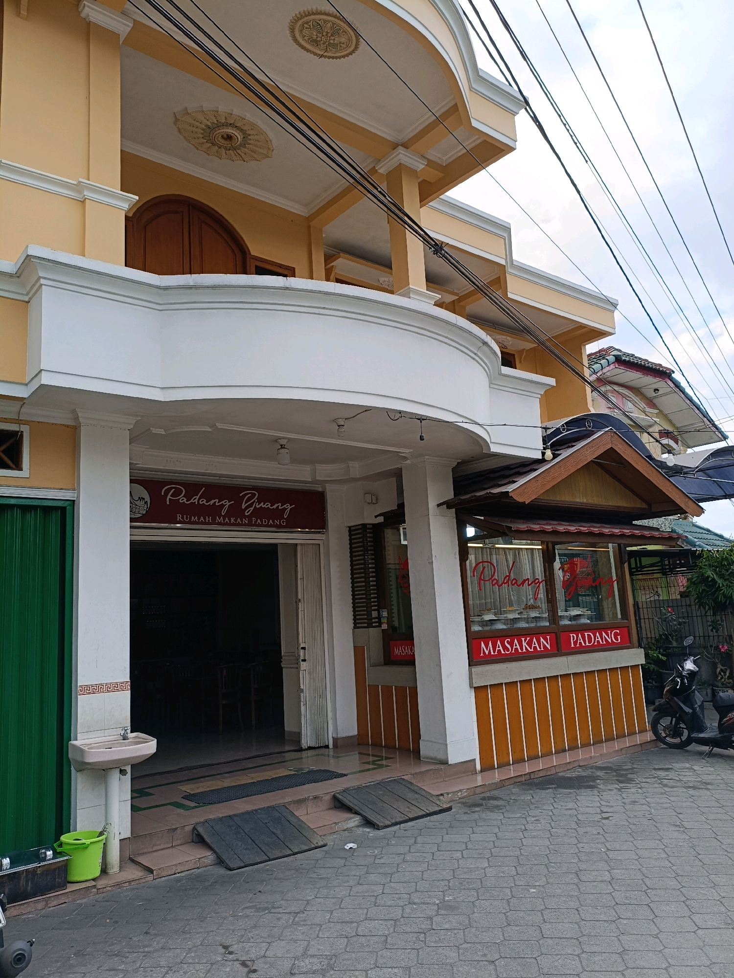 This is a ground-level, eye-level, medium shot of the exterior of a Padang restaurant named "Padang Buan." The restaurant is a two-story building painted yellow with white trim. The ground floor features a large open entrance with a maroon sign above it that reads "Padang Buan RUMAH MAKAN PADANG." To the right of the entrance is a smaller display area with windows showcasing food and a maroon sign that reads "MASAKAN PADANG." A dark-colored scooter is parked to the right of the display area. The ground in front of the restaurant is paved with gray cobblestones. A green metal door is visible to the left of the entrance, and a white sink with a green bucket is positioned next to it. Power lines are visible overhead against a slightly overcast sky. The scene is located in Ngaglik, Indonesia.