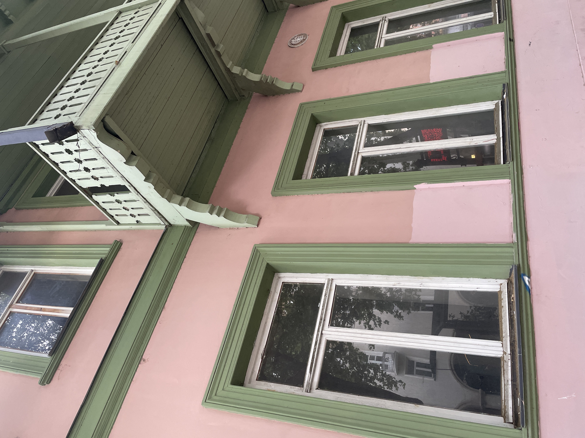 The image shows a close-up, low-angle exterior view of a pink building with green trim and a green balcony. The building has at least three windows visible, each with white frames and panes that reflect trees and another building. The balcony has a decorative wooden railing and supports. The building appears to be located in the city of Tbilisi, Georgia, judging by the architectural style which is typical of older buildings in the city. There are no people visible in the image, and no actions or interactions are depicted. The environment is urban, with the focus solely on the architectural features of the building.