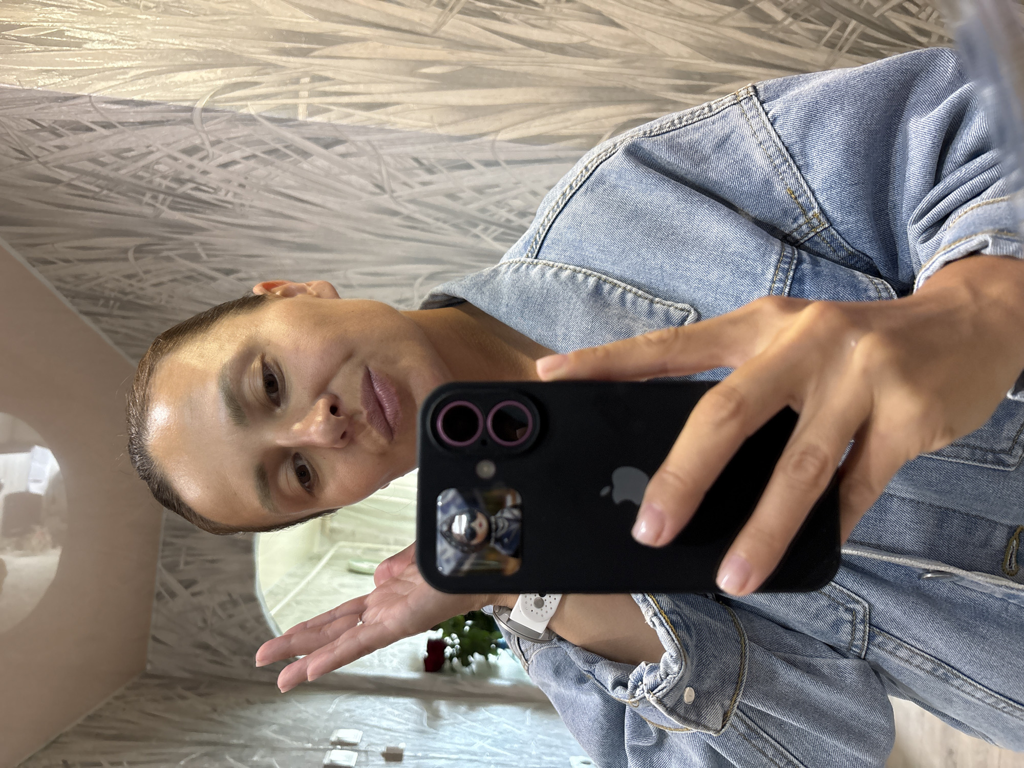 A woman wearing a light blue denim jacket takes a selfie with a black smartphone in a bathroom. The bathroom has marble-patterned walls, a white sink, and white square accessories. A floral arrangement with red roses is visible in the reflection on the phone's camera. The exact location is the city of Chaikovsky, Russia.