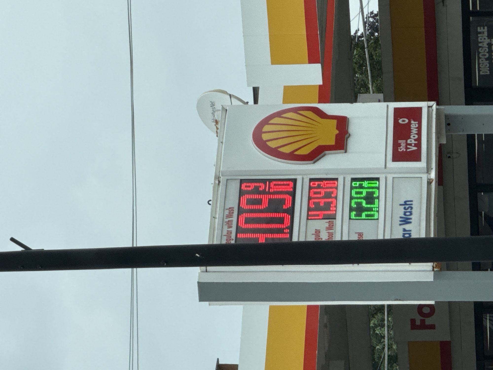 A gas station sign displays fuel prices on a cloudy day in Lincolnwood, Illinois, United States of America. The sign, featuring the Shell logo, lists the price for "Regular with Wash" at $4.09, "Regular without Wash" at $4.39, and "Diesel" at $6.29. To the right of the fuel prices, "Shell V-Power" is advertised. In the background, the yellow and red stripes of the gas station's canopy are visible, along with a satellite dish labeled "HughesNet". A utility pole is positioned in the foreground, partially obscuring the sign.