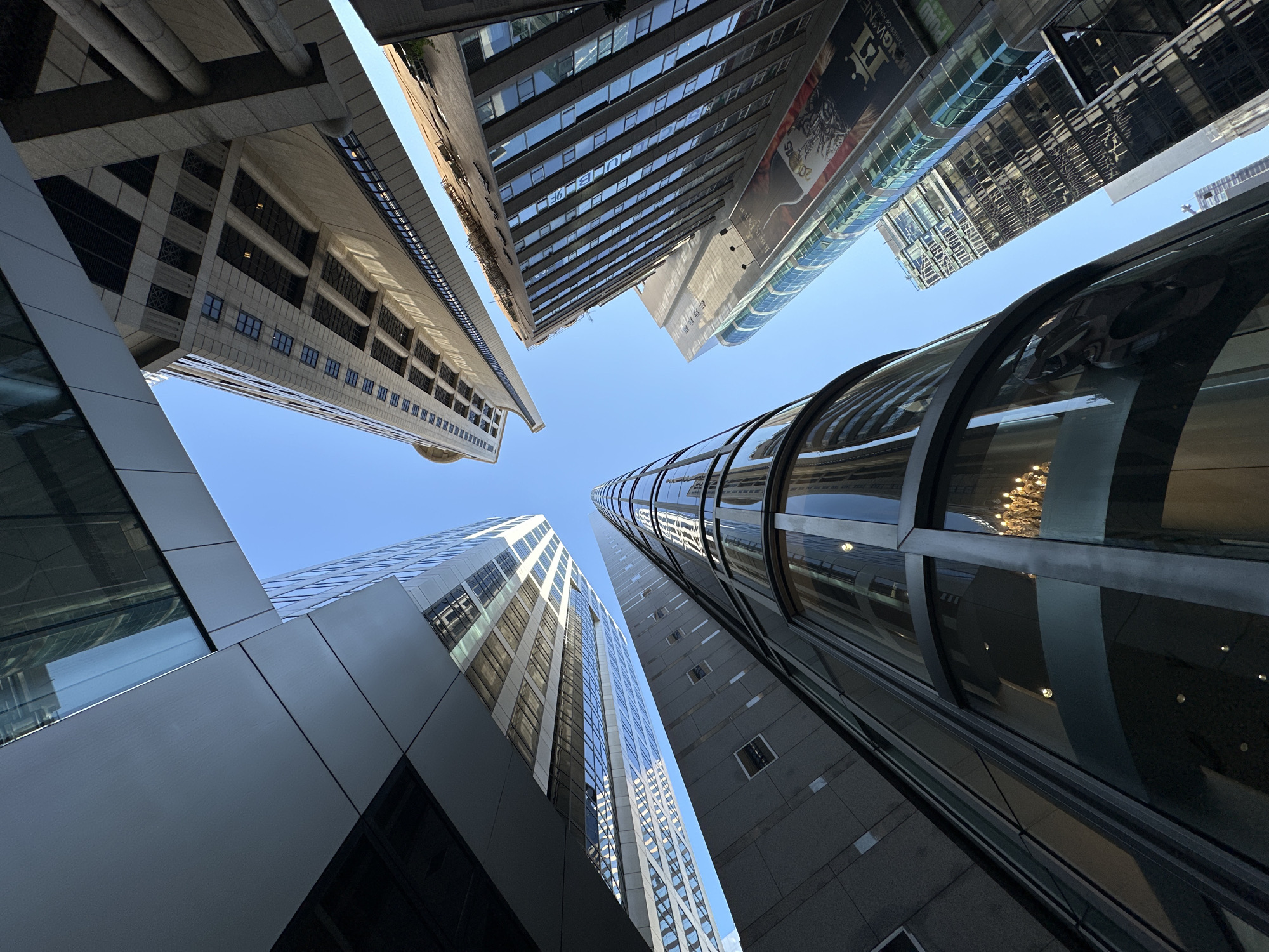 An upward-angle shot of several modern skyscrapers in Hong Kong, China, against a clear blue sky. The buildings exhibit a variety of architectural styles, with glass facades, metallic elements, and concrete structures. One building, on the far left, features a sleek, metallic finish with a glass-covered section reflecting the sky and adjacent buildings. To its right, a taller building with a more traditional design has rows of windows and visible structural beams. The central focus is a curved glass skyscraper with an ornate chandelier visible through its lower levels. Further to the right, another skyscraper displays a dark glass facade with vertical accents, and a large banner advertising "MANGINES" and "ROBER PATER" is prominent. The buildings are closely clustered, creating a sense of urban density. The lighting suggests it is daytime, with sunlight reflecting off the glass surfaces. There are no people visible in the frame.