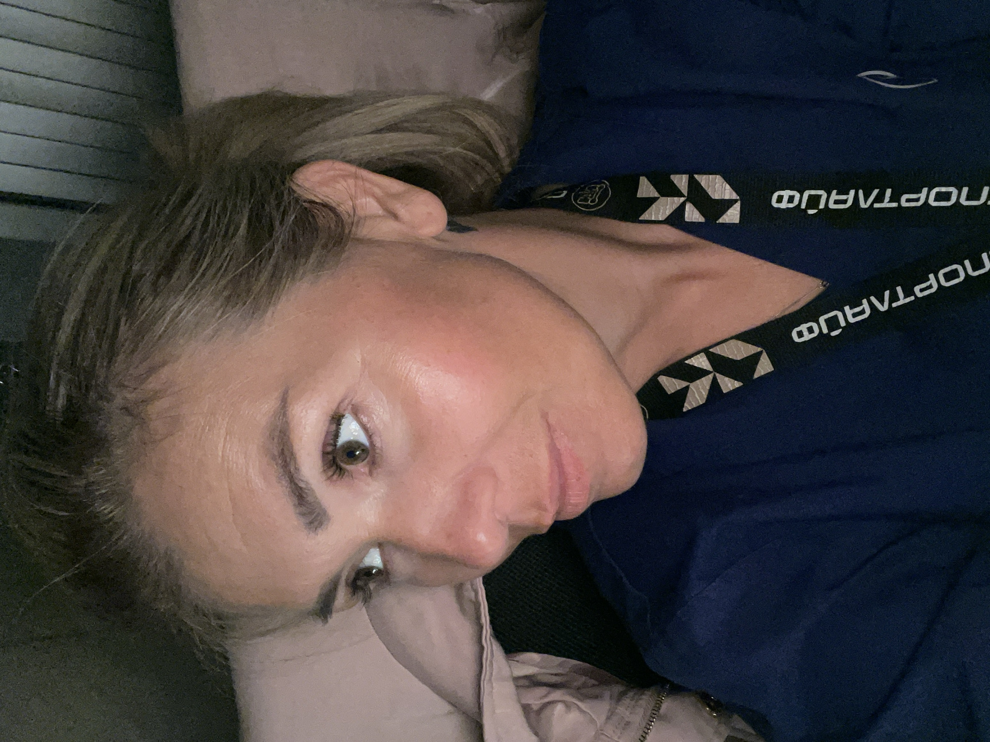 A close-up, sideways portrait of a woman lying down. She has blonde hair pulled back, and her head is tilted, showing her profile and right eye. The woman's skin is fair with some blush on her cheeks. Her right eye is open and looking towards the viewer, while her left eye is partially visible and looking away. She has dark eyebrows and lashes. Her lips are slightly parted. She is wearing a dark blue garment with a black band that has white lettering and a geometric pattern. The lettering appears to be "СПОРТЛАЙФ" in Cyrillic. The background is out of focus, with a textured surface that could be a wall or blinds on the left, and a soft, light-colored material, possibly bedding, on the right. The lighting appears to be artificial and is focused on the woman's face. The overall mood is relaxed, with a hint of curiosity in her gaze.