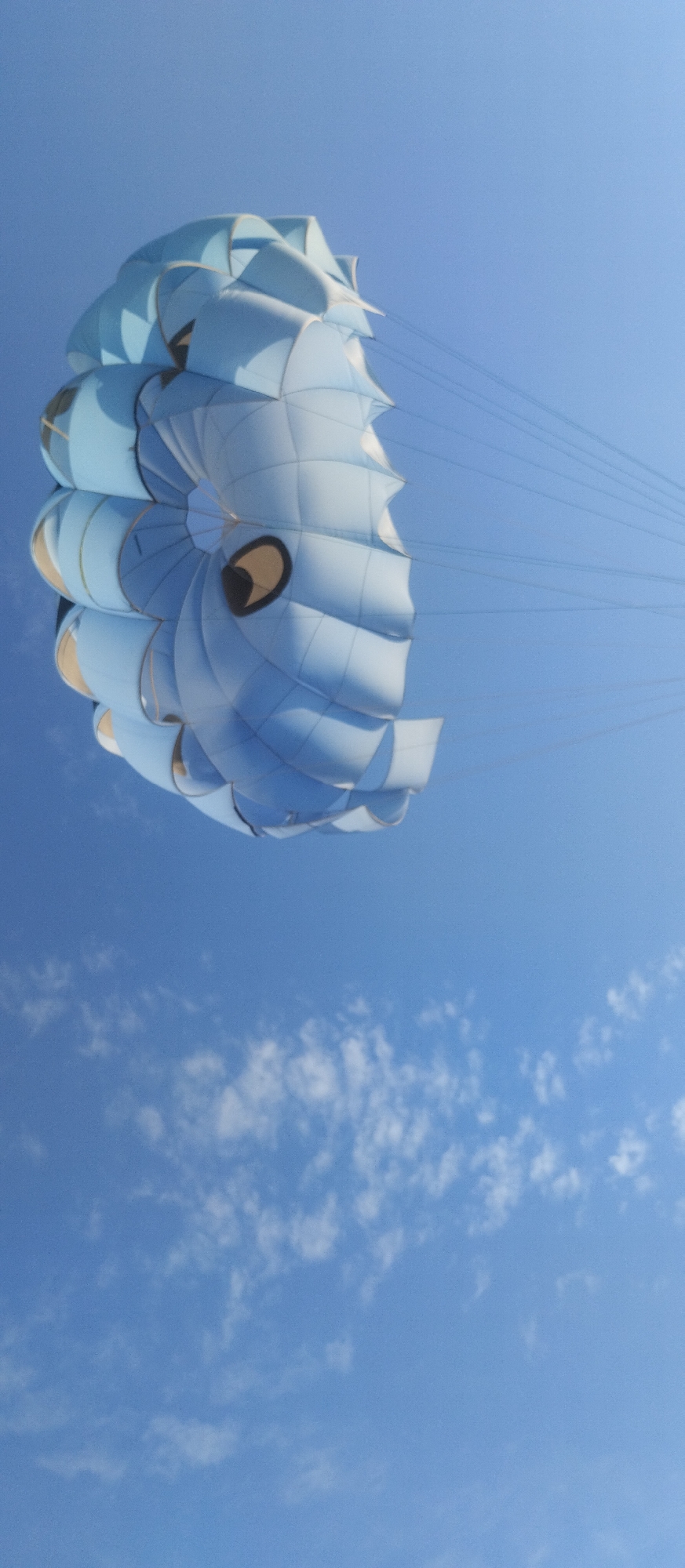 This is a low-angle, vertical, outdoor, medium shot of a parasail against a bright blue sky. The parasail is predominantly light blue and white, with a spiral pattern and two dark circular eye-like markings. It appears to be inflated and taut, suspended in the air by numerous thin, dark lines that extend upwards and to the right, suggesting it is in motion or tethered to a boat. The lower portion of the image shows scattered, wispy white clouds against the blue sky. No people or city structures are visible. The image captures a recreational activity, likely parasailing, over an open body of water, consistent with the tourist resort town of Cholpon-Ata, Kyrgyzstan, which is situated on the shores of Lake Issyk-Kul.