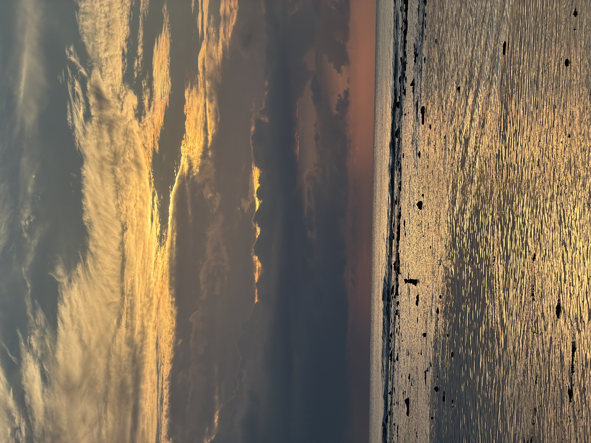 The image depicts a serene beach scene at sunset or sunrise. The sky is filled with dramatic clouds, painted in hues of orange, yellow, and gray, with streaks of sunlight breaking through. Below the sky, the calm ocean stretches to the horizon, reflecting the warm colors of the sky. The foreground shows a shallow body of water with rocks and debris scattered across the sandy bottom. The water is reflective, creating shimmering patterns of light. There are no people visible in the image, nor any specific landmarks that would indicate a particular location within "Unknown, Indonesia". The scene conveys a sense of tranquility and natural beauty.