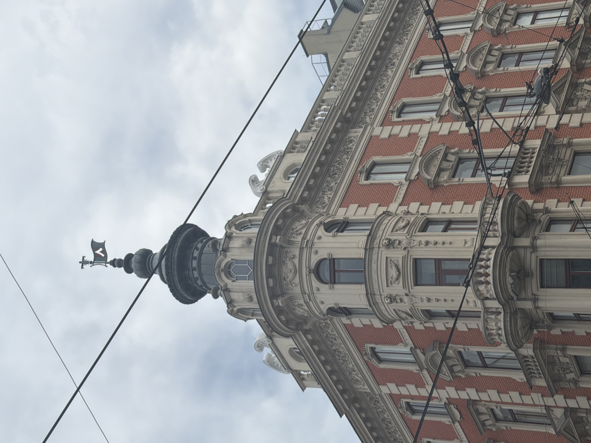 A red brick building with ornate white architectural details stands tall under a cloudy sky. The building features multiple windows with arched tops and a prominent corner turret. Atop the turret, a decorative weather vane with a flag is visible. Electrical wires crisscross the foreground, obscuring parts of the building. The architectural style suggests it is located in Vienna, Austria. No people are visible in the image.