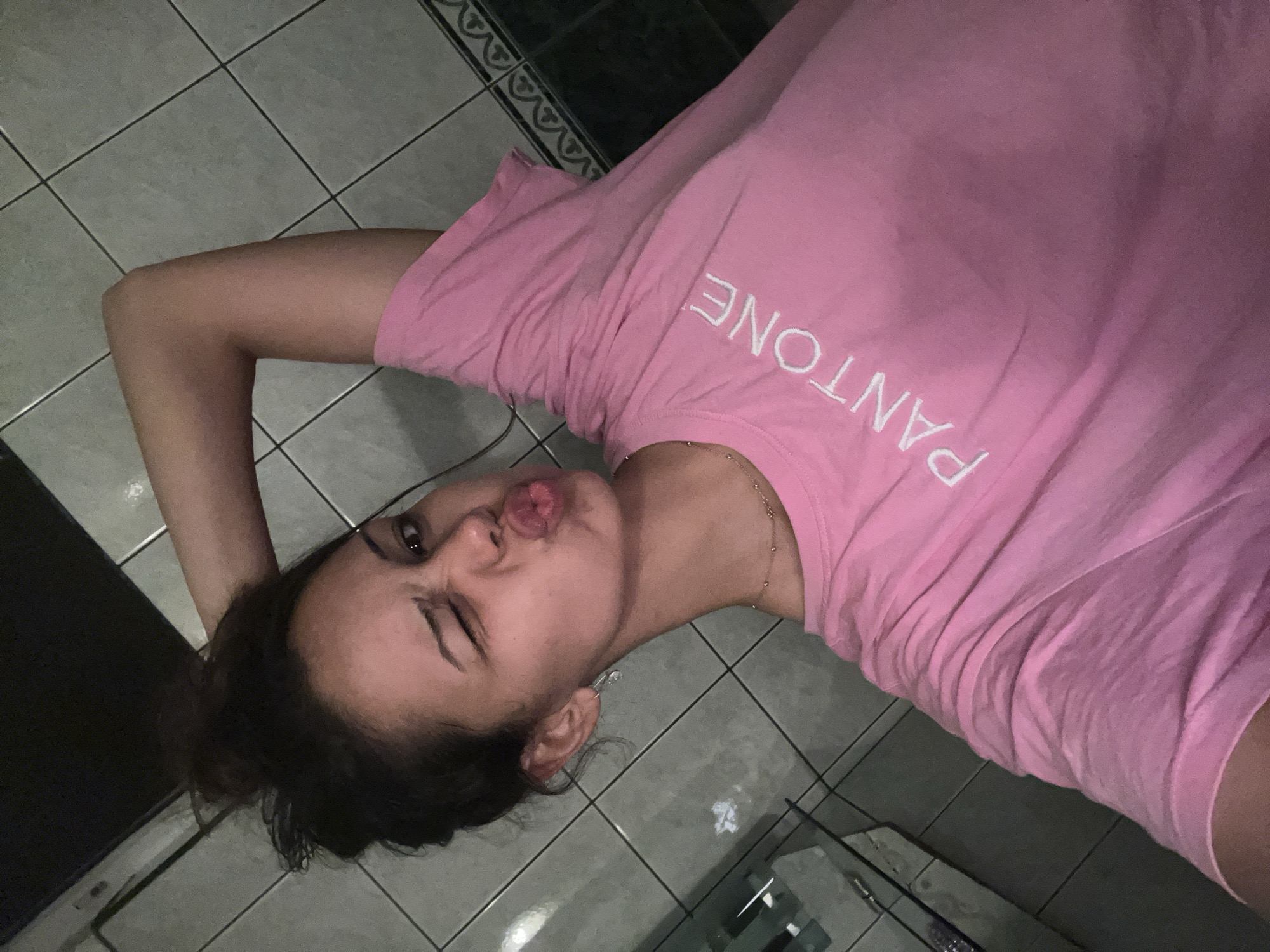 A young woman with dark hair in a messy bun is depicted in a selfie, winking her right eye and puckering her lips. Her left arm is raised, with her hand positioned near the back of her head. She wears a light pink short-sleeved t-shirt with "PANTONE" printed diagonally in white capital letters across the chest. A delicate gold-colored chain necklace and a silver-colored safety pin earring are visible on her. The background consists of light gray square wall tiles and floor tiles. A dark, patterned border tile runs along the bottom edge of the wall, and a reflective surface, possibly a mirror or glass, is partially visible in the lower-left. The scene is illuminated by artificial light.