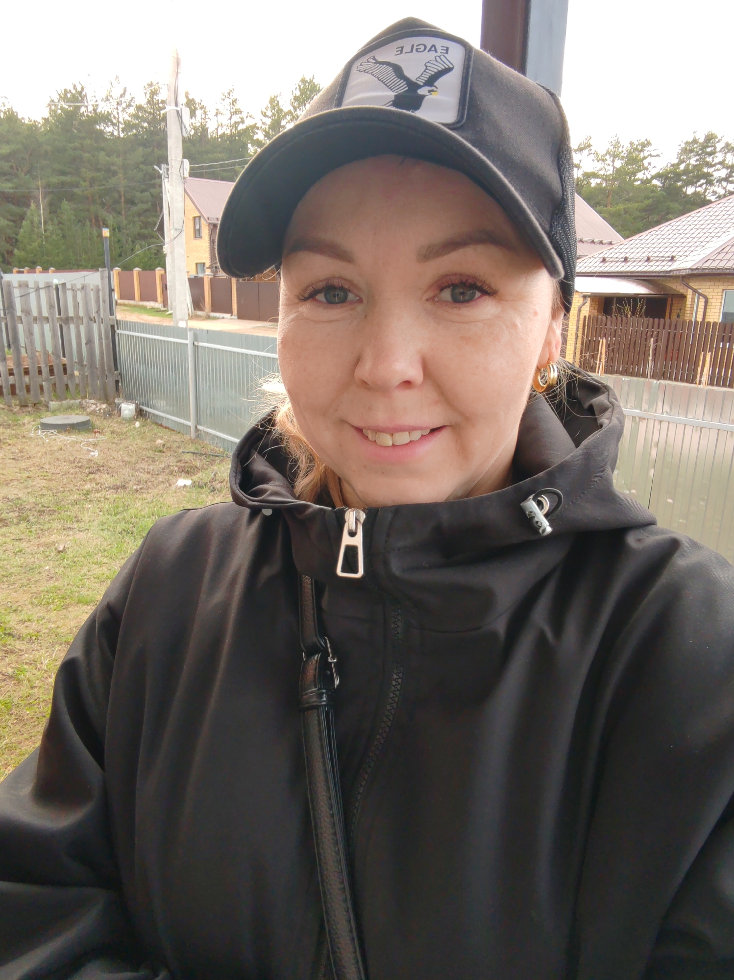 A woman is in a selfie pose outdoors in Chaikovsky, Russia. She is fair-skinned, wearing a black hooded jacket and a black baseball cap featuring an eagle logo on the front. She has blonde hair and gold hoop earrings. Behind her is a rural scene with fences, houses, and trees. A utility pole is visible in the background. The scene appears to be a residential area on a cloudy day.