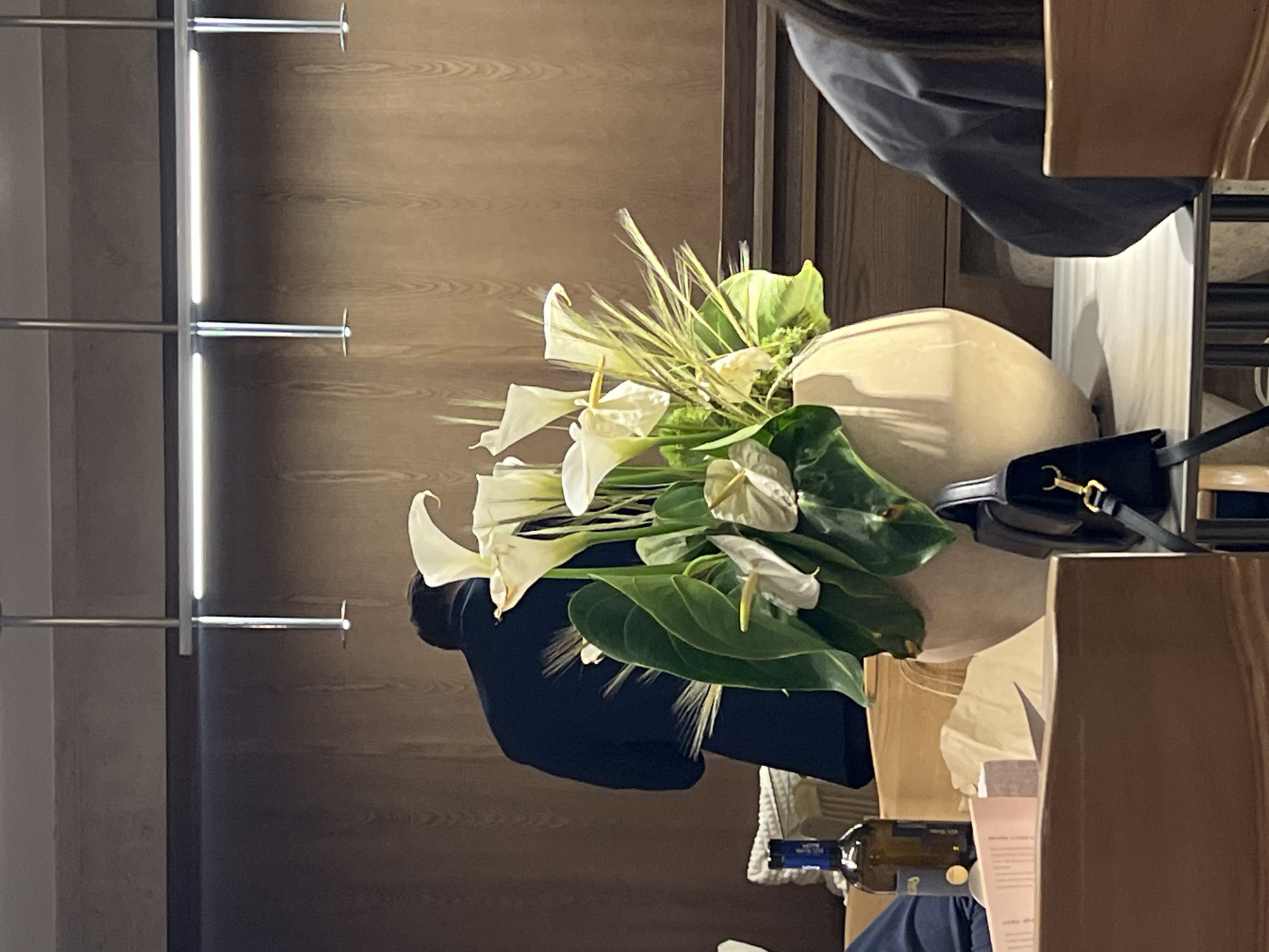 The image shows a floral arrangement in a beige vase, consisting of white lilies and green foliage. The vase sits on a surface cluttered with various items, including a dark-colored purse with a gold clasp, a wine bottle, and a pamphlet with text. A person wearing a dark-colored garment is partially visible behind the arrangement. In the background, there is a wall with a light-colored, wood-like texture and a metal rack system. The location is Unknown, Unknown.