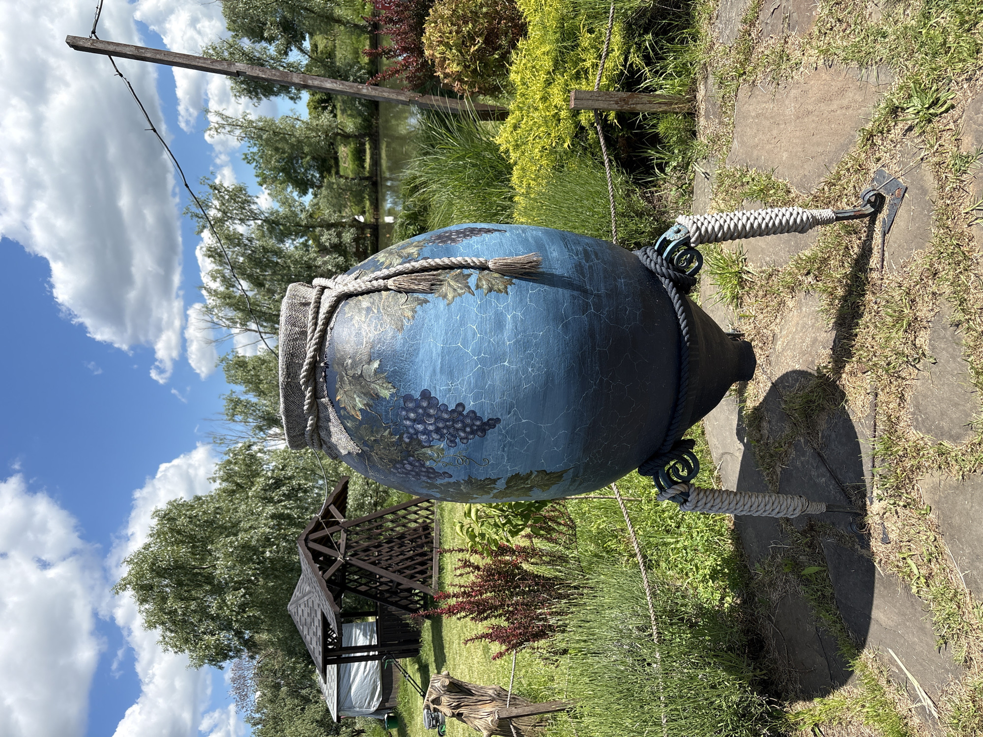 This image captures a vibrant garden scene, centered around a large, ornamental, blue amphora-like pot. The pot is decorated with painted grapevines and bunches of dark purple grapes, featuring a distressed, crackle-paint finish. A thick, braided rope with a tassel is draped over its neck, and additional ropes are wrapped around its metal handles, which are themselves coiled with a lighter, woven rope. The pot stands on a dark metal base with two curved, rope-wrapped supports.
The setting is an outdoor garden. The foreground features patchy grass and stepping stones on the right. In the midground, there's a lush variety of plants and bushes, including some with reddish-purple foliage and bright green specimens. A dark wooden gazebo or pergola structure is visible on the left side, and a small body of water, possibly a pond or lake, can be seen further back, reflecting the sky. The background is dominated by large green trees and a bright blue sky with scattered white clouds. A wooden post with a thin wire attached is also visible.
The scene is devoid of people, presenting a tranquil and decorative landscape. The clear sky and distinct shadows indicate a bright, sunny day, likely mid-day. There is no visible text in the image. The user-provided context specifies the location as Gay-Kodzor, Russia, although no specific visual cues within the image confirm this.