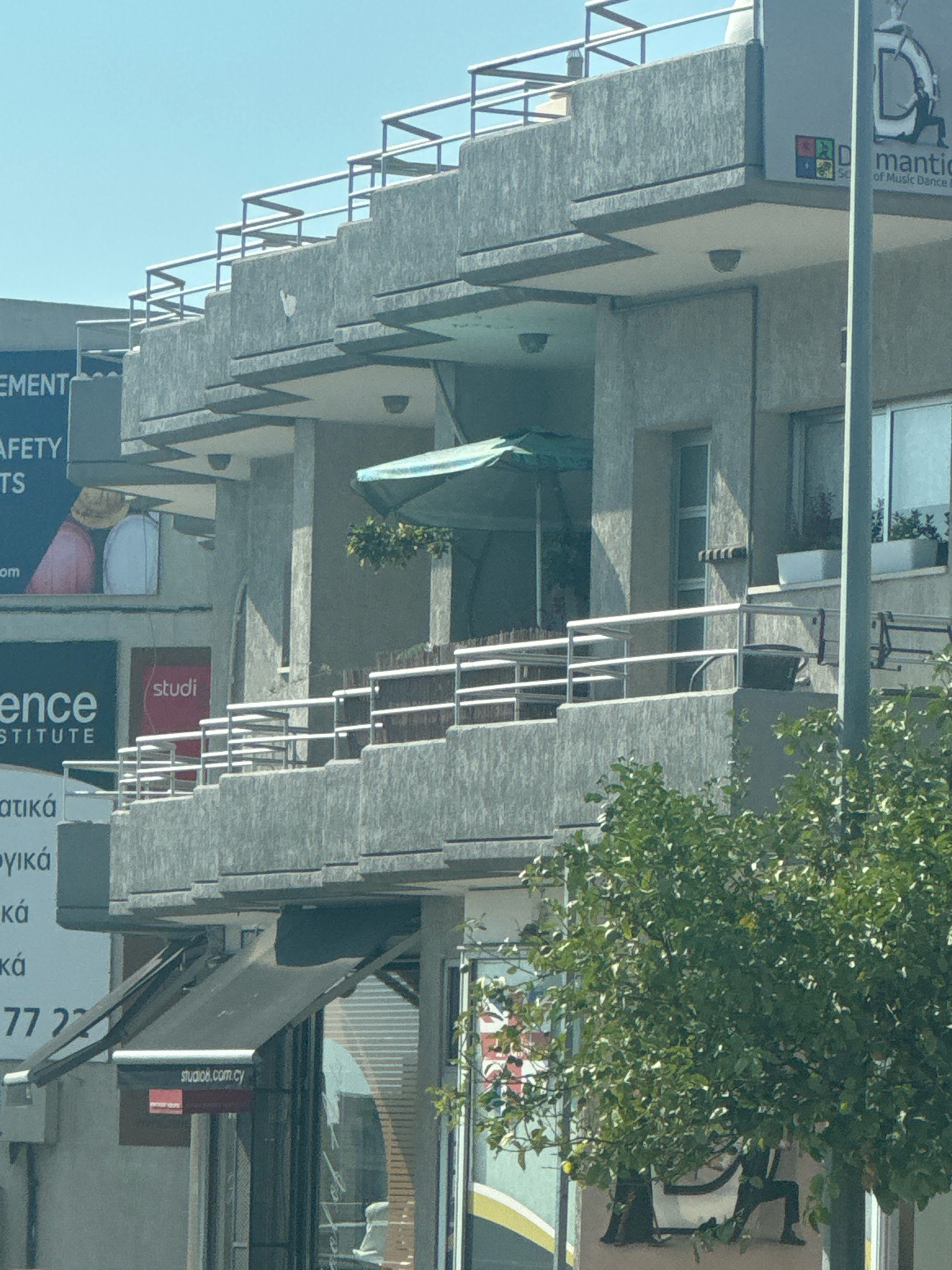The image shows a multi-story building with a concrete facade in Mesa Geitonia, Cyprus. The building features balconies with metal railings, several windows, and a few awnings over storefronts on the ground level. Signs are visible on the building, including one with text that appears to be in Greek, a "studio8.com.cy" sign, and a sign for "Mantiic of Music Dance". There are also some posters or advertisements on the side of the building. A green leafy tree is partially obscuring the lower right portion of the building. The sky is clear and blue.