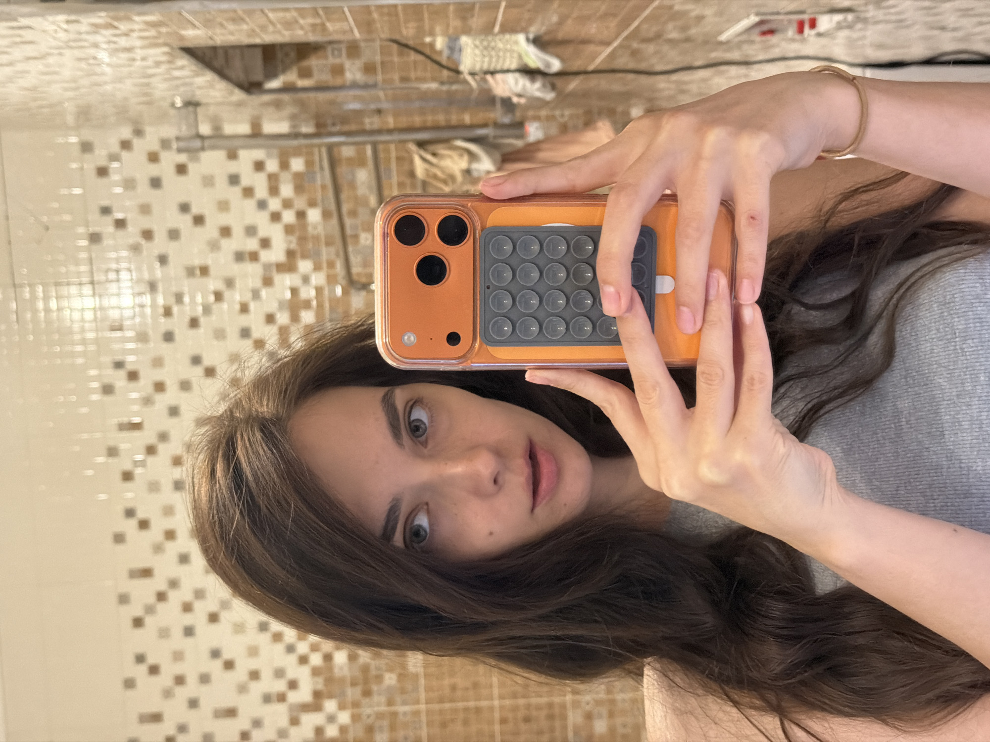 A young woman with long, dark, wavy hair is taking a selfie in a bathroom. She is wearing a gray tank top and a gold bracelet on her right wrist. She is holding an orange phone with a pop-it case in her hands. The bathroom has tiled walls with a mosaic pattern of brown and beige squares. A towel rack is visible in the background, along with some towels and a black cord.