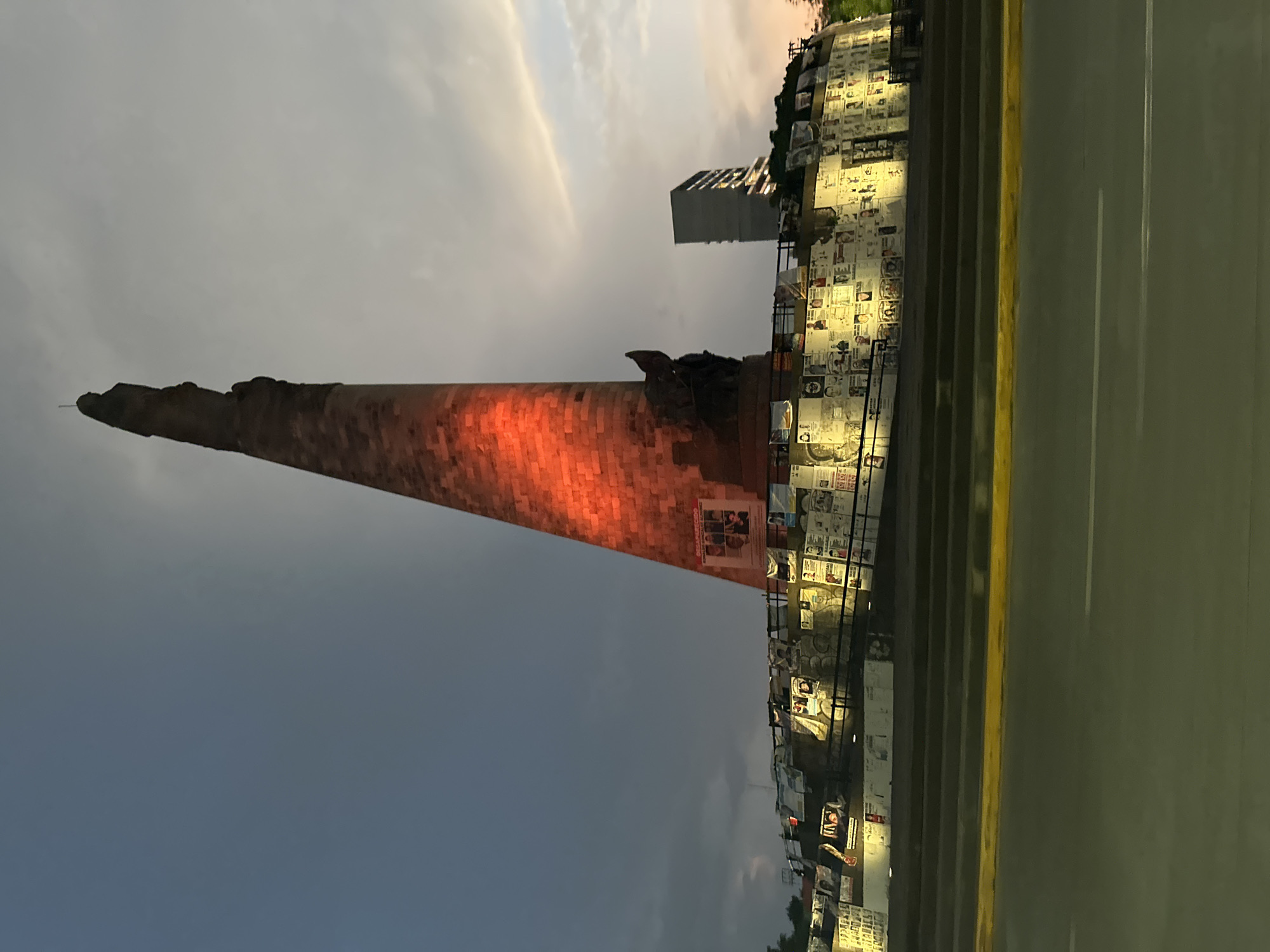 The image depicts the "Monumento a los Niños Héroes" in Guadalajara, Mexico, at dusk or night. The monument, lit with red light, is a tall obelisk-like structure. Adjacent to it is a tiered platform covered with posters and illuminated by spotlights. A building is visible in the background behind the monument. A grey road is in the foreground. The sky is partly cloudy.