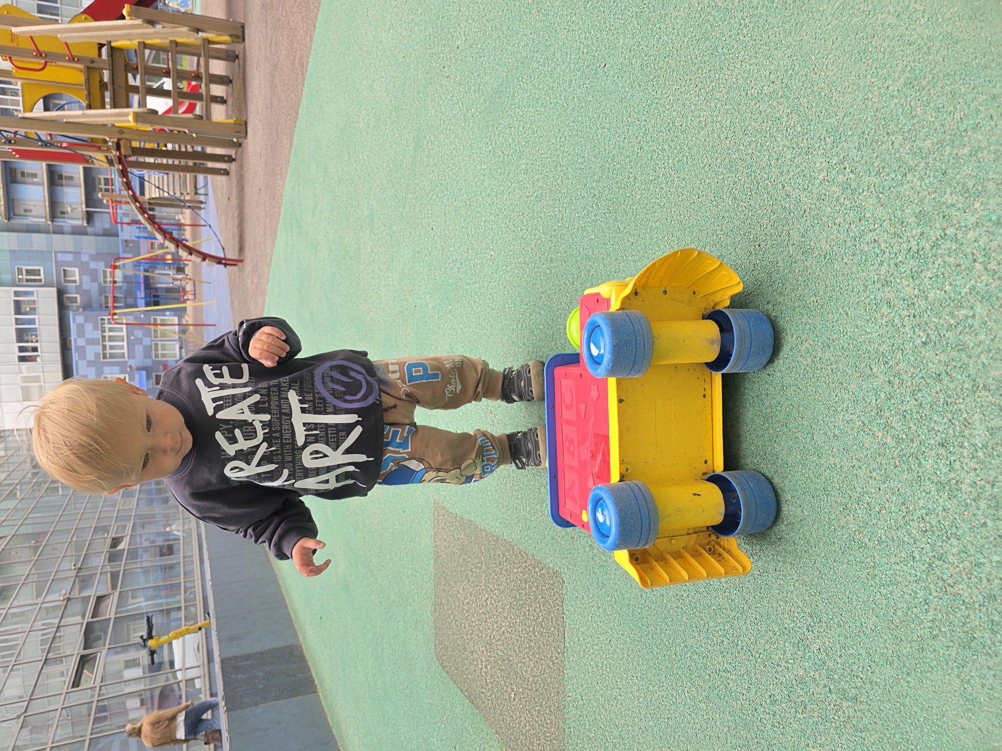 A toddler with blond hair stands on a green rubberized playground surface, looking down at a overturned yellow and red toy truck. The child is wearing a black sweatshirt with white lettering that says "CREATE ART" and brown pants with cartoon characters and the letter "P". The playground is located in front of a modern residential building with a glass facade. In the background, there are playground structures, including swings and a wooden climbing frame. A woman in a brown jacket and blue jeans is visible walking in the distance. The scene appears to be in the city of Saint Petersburg, Russia.