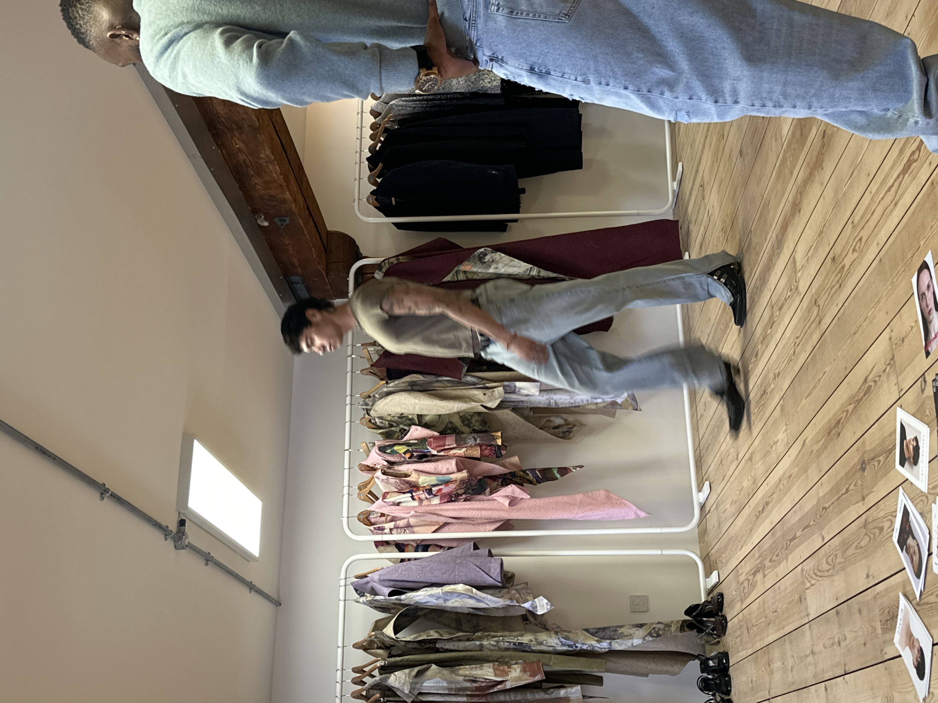 The image captures an indoor scene, likely a creative studio, showroom, or retail space, with two individuals and multiple clothing racks. The perspective is from a slightly elevated angle, looking down.
In the upper portion of the frame, a person with short dark hair is partially visible. They are wearing a light grey long-sleeved sweatshirt and light blue jeans. A watch with a dark strap and a metallic case is visible on their left wrist. This person appears to be standing or leaning over, possibly inspecting items on a clothing rack that is partly obscured by their body.
In the middle-right area, another individual is captured mid-movement, appearing to be walking or possibly jumping with their legs extended. They are wearing a light brown or khaki short-sleeved t-shirt, light blue jeans, and black shoes. Their dark hair is styled in a way that suggests a mullet. Their body is slightly blurred, indicating motion. This person is positioned between two white, minimalist clothing racks.
The room features light-colored walls, possibly off-white or cream. A large wooden beam runs across the upper part of the wall, suggesting structural elements of an older building. A metal pipe runs horizontally along the left wall, with a rectangular white light fixture emitting a bright glow attached to it. The floor is made of light-colored wooden planks, arranged in a diagonal pattern.
Two primary clothing racks are visible. The top rack, partly obscured, holds dark-colored garments, possibly trousers or skirts, on light wooden hangers. The lower racks are filled with numerous garments, many of which appear to be made from patterned fabrics. There are items in shades of pink, purple, and various earthy tones with intricate patterns. These clothes are folded or draped over wooden hangers, creating stacks of fabric.
On the wooden floor in the lower right section of the image, several small rectangular printouts or photographs are scattered. These appear to be portraits of individuals, possibly models or design inspirations. One clearly visible print shows a woman's face with text partially visible below it, which appears to read "MATTHEW" or a similar name. Other prints show a person with dark hair looking downwards and another face. Some dark-colored shoes or sandals are also visible on the floor near the bottom clothing rack.