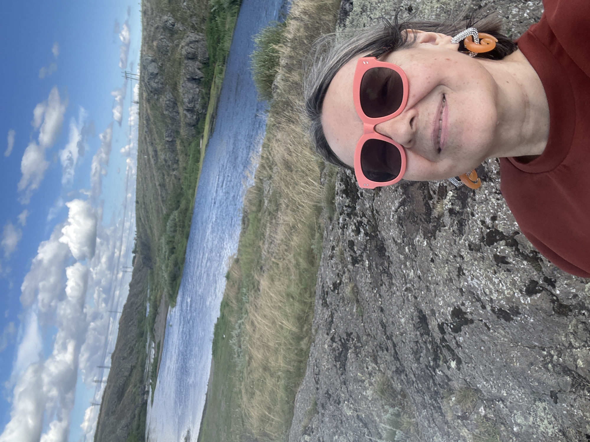 In a selfie taken outdoors, a woman with graying hair and wearing pink sunglasses and orange hoop earrings smiles at the camera. She has a maroon shirt on. The background depicts a natural landscape with a wide, blue river flowing through a valley. The riverbanks are lined with dry, golden grasses and green vegetation. Beyond the river, rolling hills covered in sparse vegetation and rocky outcrops rise towards a sky with scattered white clouds. Power lines and poles are visible in the distance on the hills, suggesting proximity to some form of infrastructure. The foreground shows rough, textured grey rocks.