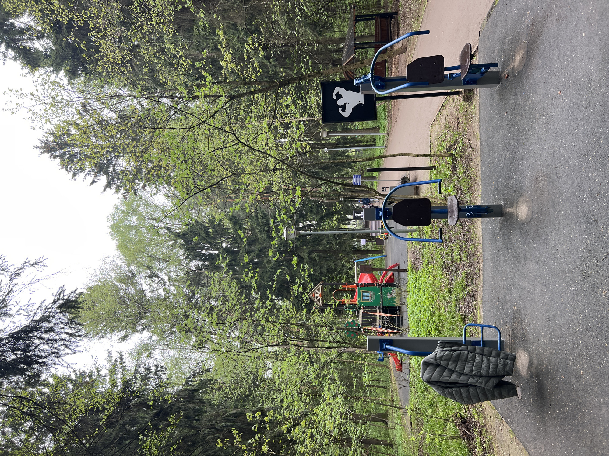 The image shows a park scene in Vnukovo, Russia. The foreground features exercise equipment with blue metal frames and black seats on a paved path. A green quilted jacket is draped over one of the pieces of equipment. Further into the scene, there is a sign with a white figure flexing muscles on a black background. A green and red playground structure is visible amidst trees. A walking path extends into the background, with additional park benches and possibly other people visible. Trees surround the open area, and the sky is overcast.